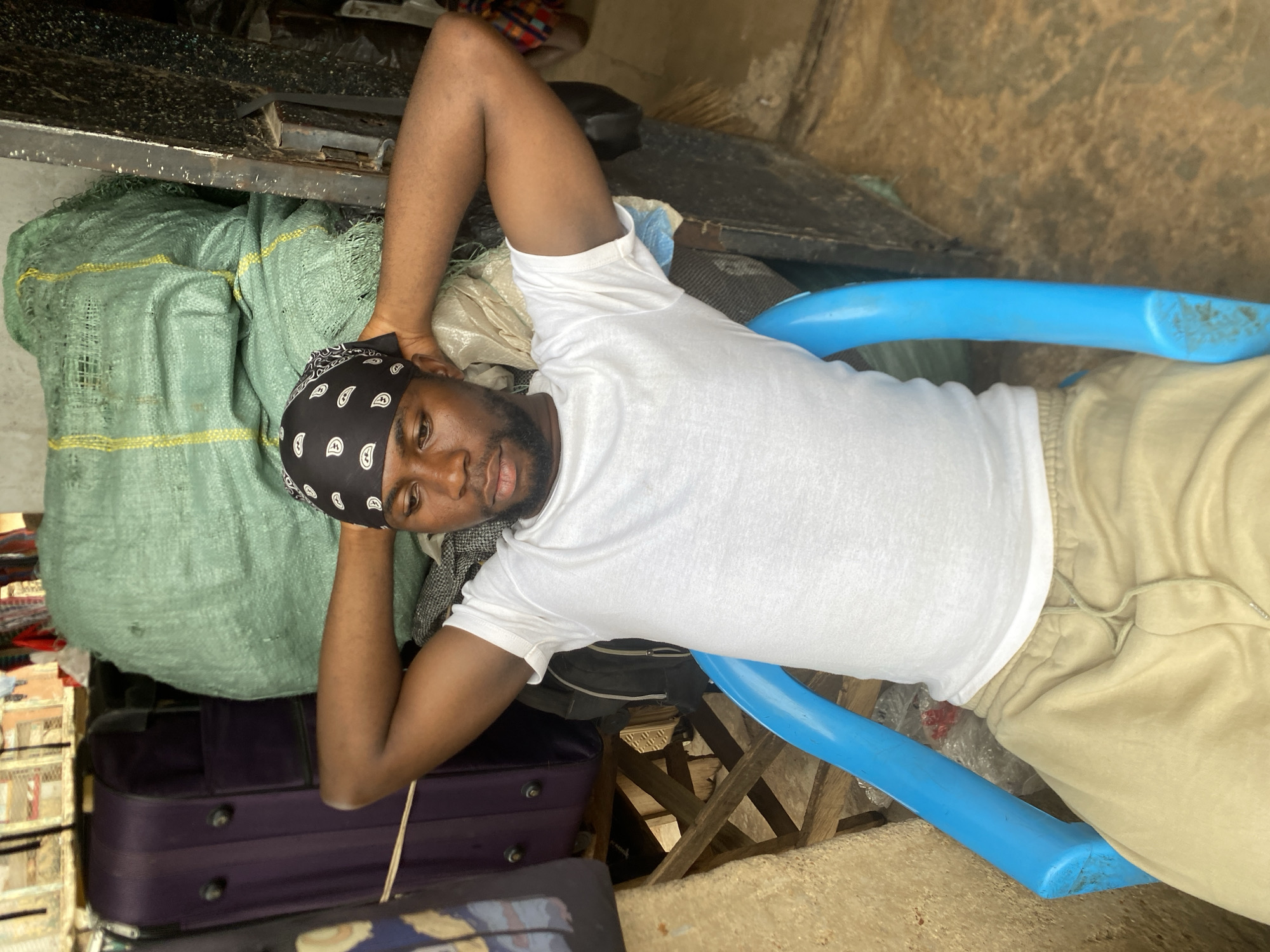 A man is lying on his back on a blue plastic chair in a crowded indoor space. The man is wearing a white t-shirt, khaki pants, and a black bandana with white paisley print. His arms are raised, hands behind his head. The chair is situated amidst luggage, bags, and other assorted objects. The location is Bukuru, Nigeria. The area appears to be enclosed with concrete walls.