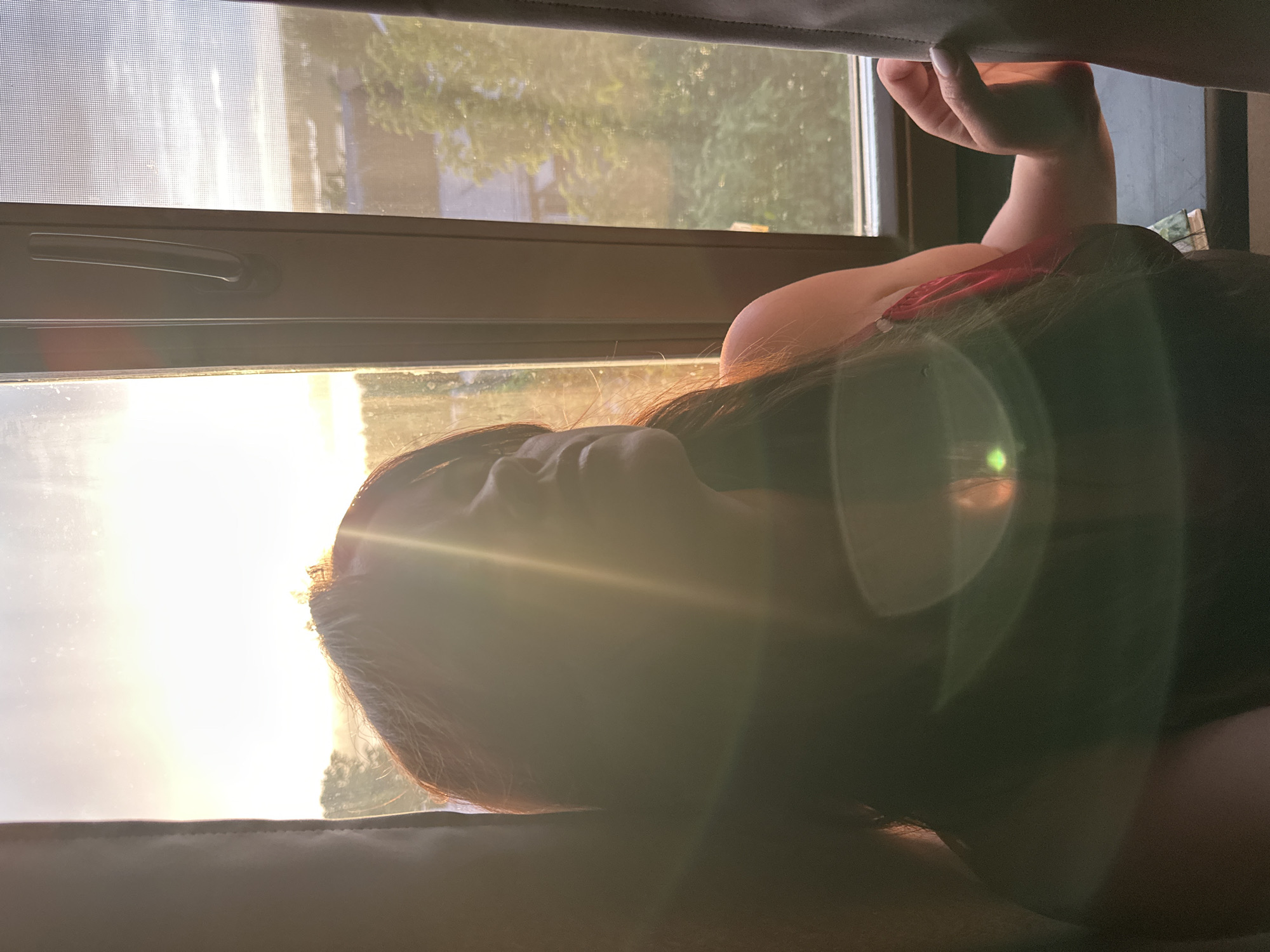 An image captures a person resting indoors by a window, bathed in warm, golden sunlight, located in Chaikovsky, Russia. The original image is oriented 90 degrees counter-clockwise.
The primary subject is a person, likely female, with long, dark hair, positioned with their head to the left and body extending to the right. Their profile is visible, looking out through the window. Bright sunlight and significant lens flare partially obscure their face and upper body. A hand, with white-painted fingernails, is visible reaching upwards, and a hint of red fabric from clothing can be seen near the shoulder.
Through the window, a serene outdoor scene is visible. Lush green foliage, indicative of trees, and what appears to be a distant building can be seen. Details of the window itself include a screen texture on the upper pane and a dark-colored handle on the frame.
The intense, low-angle sunlight, characterized by prominent lens flares, strongly suggests that the photo was taken during either sunrise or sunset, commonly known as the golden hour. The overall atmosphere is tranquil and contemplative. No visible text is present in the image.