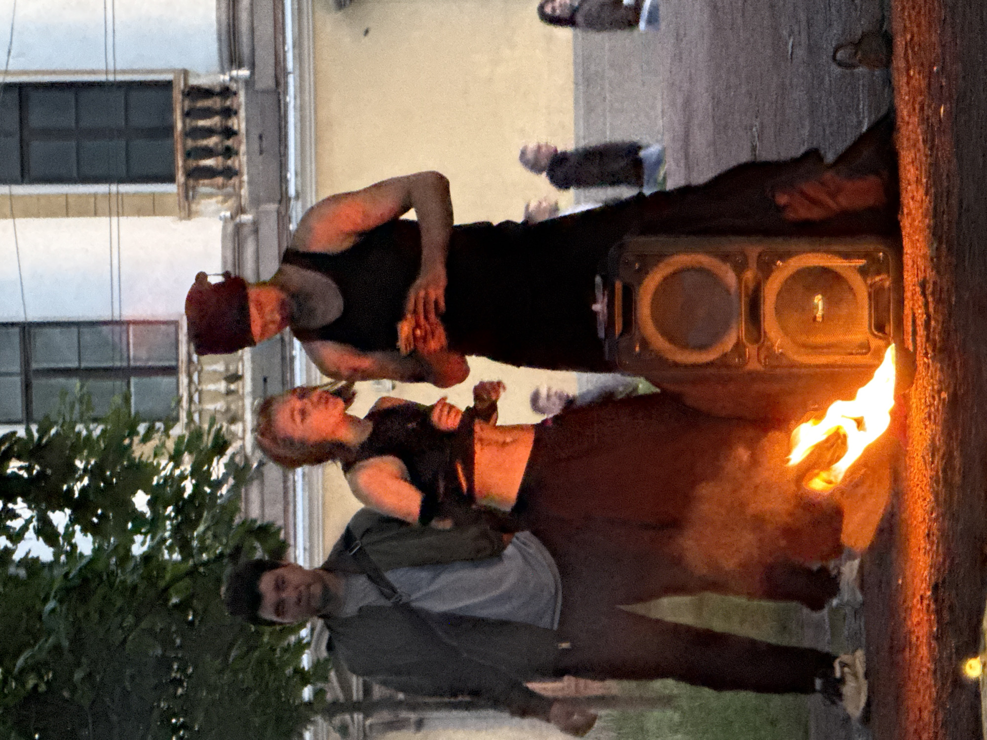 The image depicts an outdoor scene, likely in Saint Petersburg, Russia, given the architectural style of the building in the background. Three individuals are the primary subjects, gathered around a fire pit with a portable speaker. A man, wearing a dark tank top and a red beanie, stands in the center, holding an object in his hands, possibly food or a prop. To his left, a woman in a black, revealing top and a long, dark skirt is looking at him, her body angled towards the fire. To the far left, a man in a dark jacket over a grey t-shirt stands observing the scene. The fire is actively burning, casting an orange glow on the people and the speaker. In the background, a building with large windows and ornate balconies suggests a historical or cultural district. Other people are visible in the distance, appearing as spectators. The ground is paved, and there is some vegetation on the left side of the frame. The overall atmosphere suggests a performance or gathering, possibly a street performance due to the presence of the fire, speaker, and the engaged audience.