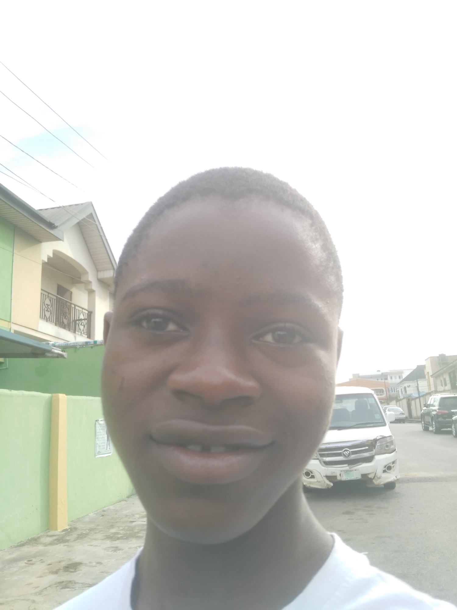 A close-up of a person with short dark hair is centered in the frame, looking directly at the camera with a slight smile. Behind them, on the left, is a green and yellow wall adjacent to a building with a beige and white facade and a balcony. To the right and further back, a white van with front-end damage is parked on a street. Other vehicles are also parked along the street in the distance. The scene appears to be in an urban residential area. The image is somewhat overexposed.