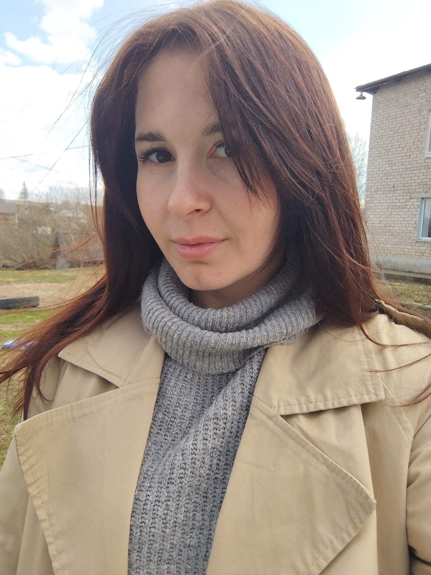 A close-up portrait captures a woman outdoors in Фоки, Russia, looking directly at the camera with a neutral to slightly contemplative expression. The subject, a woman appearing to be in her 20s or 30s, has long, dark reddish-brown hair, some strands of which are subtly windblown, and dark eyes.
She is dressed in a light beige or khaki trench coat worn over a thick, light grey knitted turtleneck sweater, suggesting cool weather. The detailed texture of the sweater is visible, providing a soft contrast to the smoother fabric of the coat.
The background reveals a bright, clear daytime sky with scattered white clouds on the left. Distant bare trees are visible, indicating either late winter or early spring. On the right, part of a light-colored brick building with a window and a dark object (possibly a light fixture or vent) near the roofline is present. The ground appears to be a mix of grass and dirt. The overall lighting is bright and natural, casting soft shadows. There is no visible text in the image.