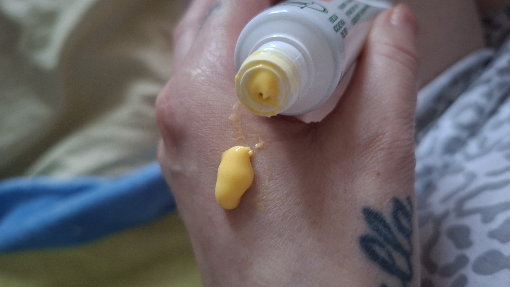 In a close-up shot, a person's hand with a tattoo on the wrist is shown. A dollop of yellow cream is squeezed onto the palm of the hand, and another smaller amount is visible on the opened tube of the cream. The background consists of light-colored fabric and a blurred blue and yellow fabric. The scene appears to be indoors, possibly in a bedroom or living area. The specific location in Забалоцкі сельскі Савет, Belarus is not discernible from the image.