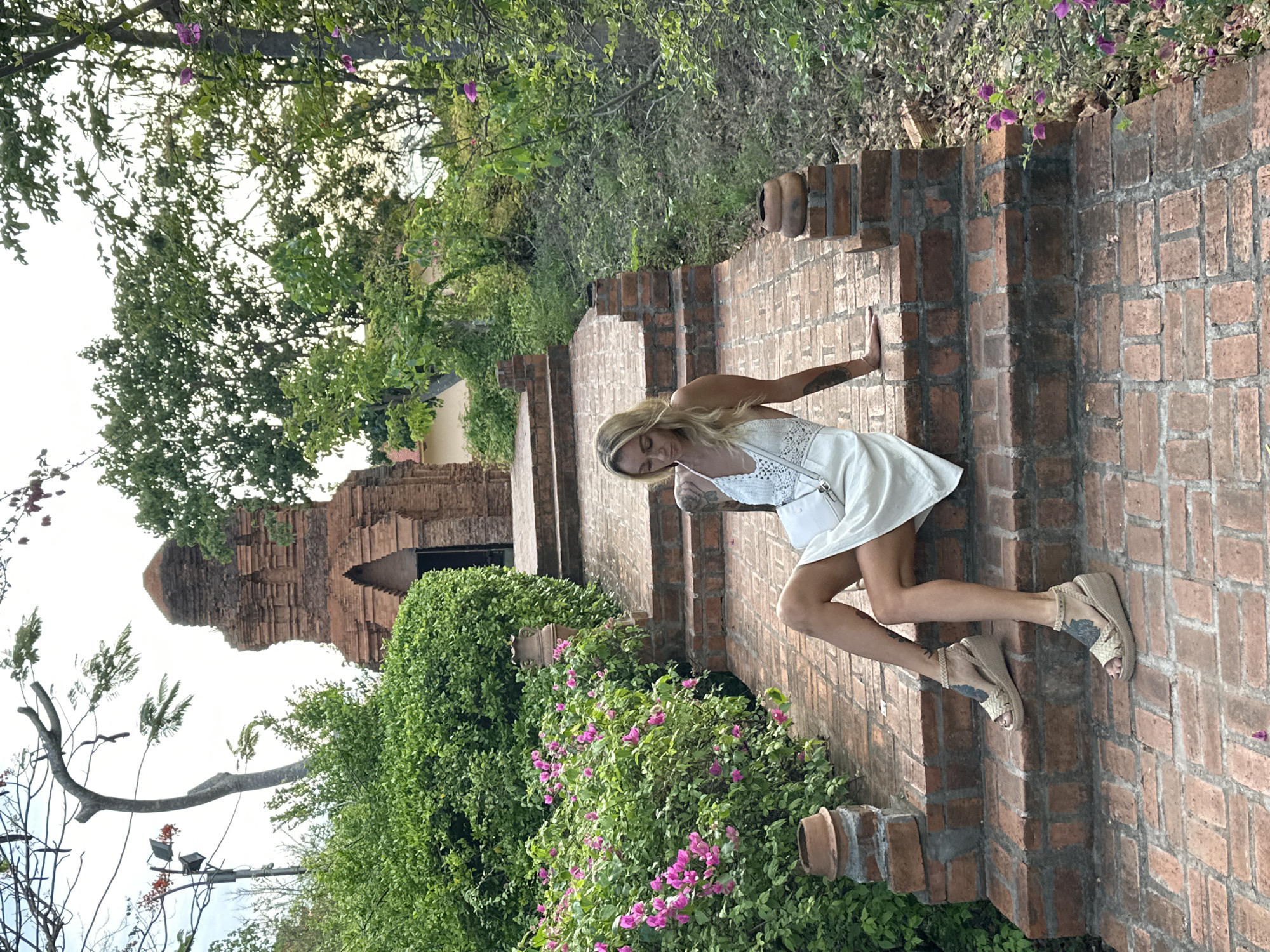 A fair-skinned woman with blond hair is walking down a brick staircase in Phu Thuy Ward, Vietnam. She wears a white dress and light-colored wedge sandals. Her left arm rests on the brick wall of the staircase. The staircase leads toward a brick building with a tower-like structure in the background, nestled among trees and greenery. The staircase is flanked by green bushes with pink flowers. A bare tree branch and light posts are visible in the background.