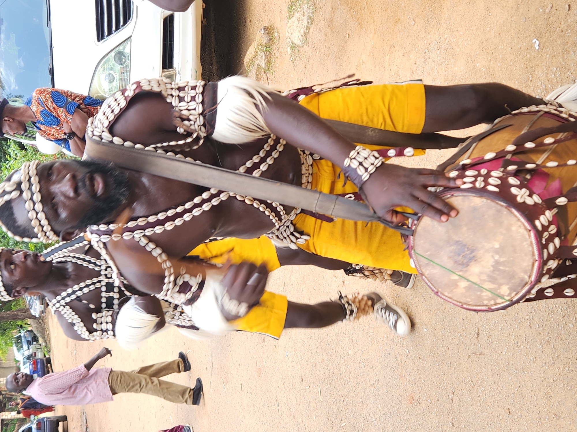 Two men in traditional attire, adorned with necklaces and bracelets made of cowrie shells, are performing a dance. One man is playing a drum, while the other stands beside him, also decorated with shell ornaments. Both are wearing yellow shorts and white loincloths. The drum is round with a reddish-brown border decorated with shells and what appear to be small pink pieces of fabric. The men have dark skin and appear to be in motion, suggesting a lively performance.
In the background, a white SUV is parked, and several other vehicles are visible further away. The ground is sandy, and some greenery can be seen to the left, hinting at an outdoor setting. The lighting suggests it is daytime, possibly late morning or early afternoon, with the sun casting some shadows. The overall atmosphere appears energetic and celebratory, indicative of a cultural event or performance. A third man in a pink shirt and brown trousers is walking in the foreground, observing the performers. The scene is set in Jos, Nigeria. No visible text is present in the image.