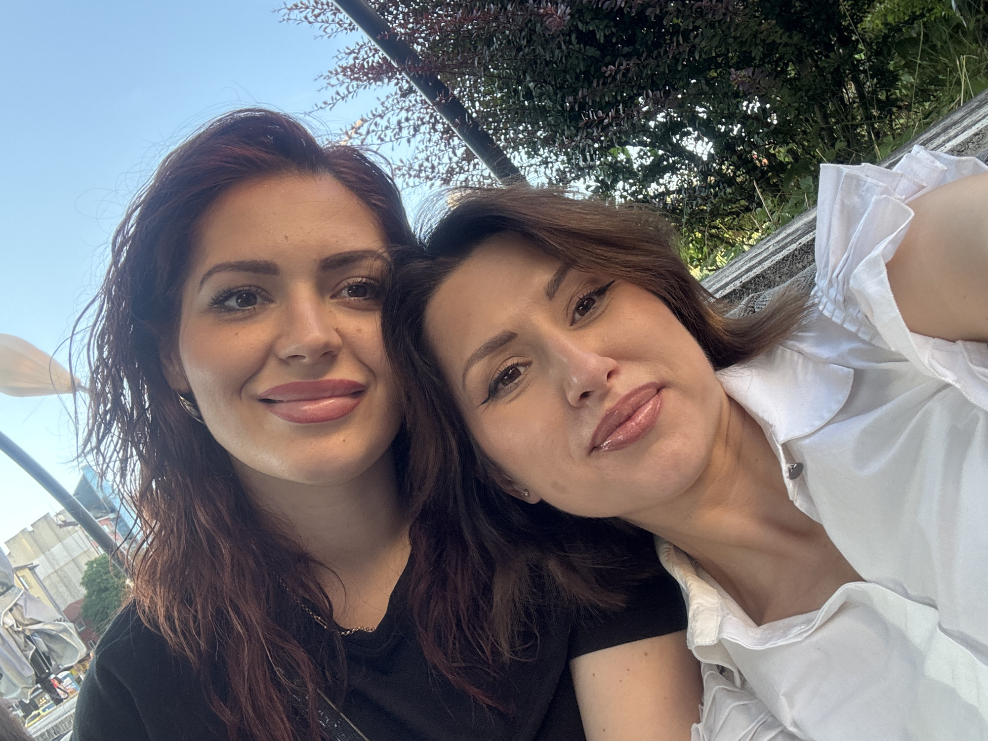 Two women are taking a selfie outdoors. The woman in the foreground is wearing a white collared shirt with ruffled sleeves and is looking at the camera with a slight smile. The woman behind her is wearing a black top and her face is partially obscured by her dark hair. Both women have makeup on. The background shows foliage and the sky. The overall setting appears to be a park or outdoor seating area in a city.
