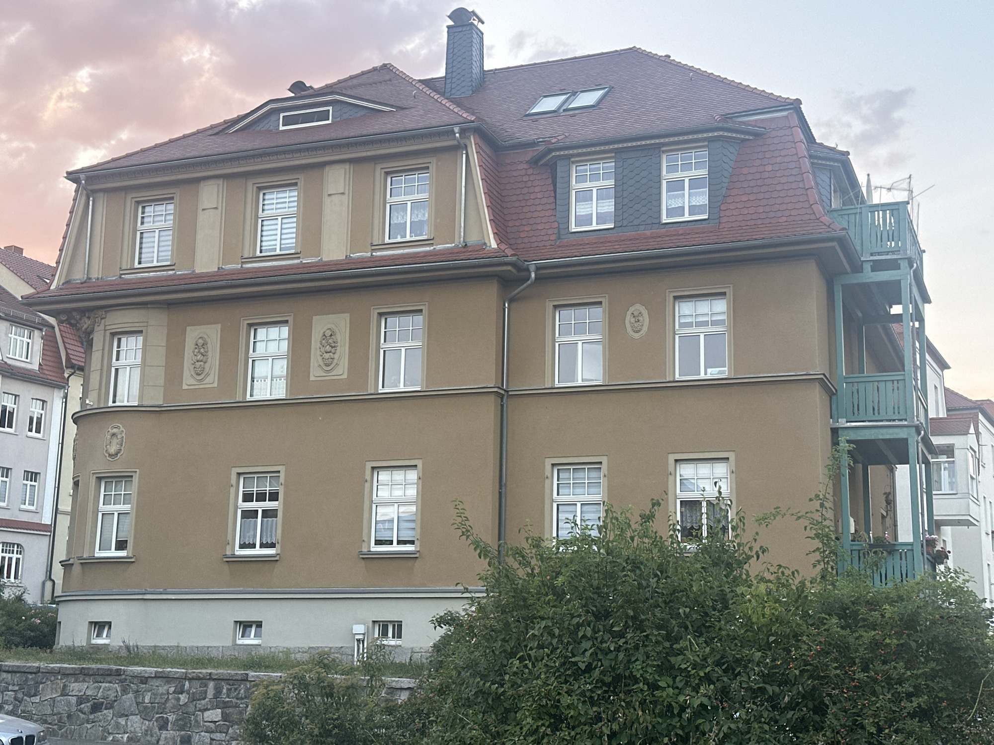 The image shows a large, multi-story building in Bautzen, Germany, under a cloudy, pinkish sky. The building has a tan facade with a red tiled roof. There are numerous windows on each level, many with white frames and blinds. Decorative elements, such as oval reliefs, are visible on the facade. A green, multi-level balcony extends from the right side of the building. In the foreground, there is a stone wall and dense green foliage. The scene appears to be a residential area.