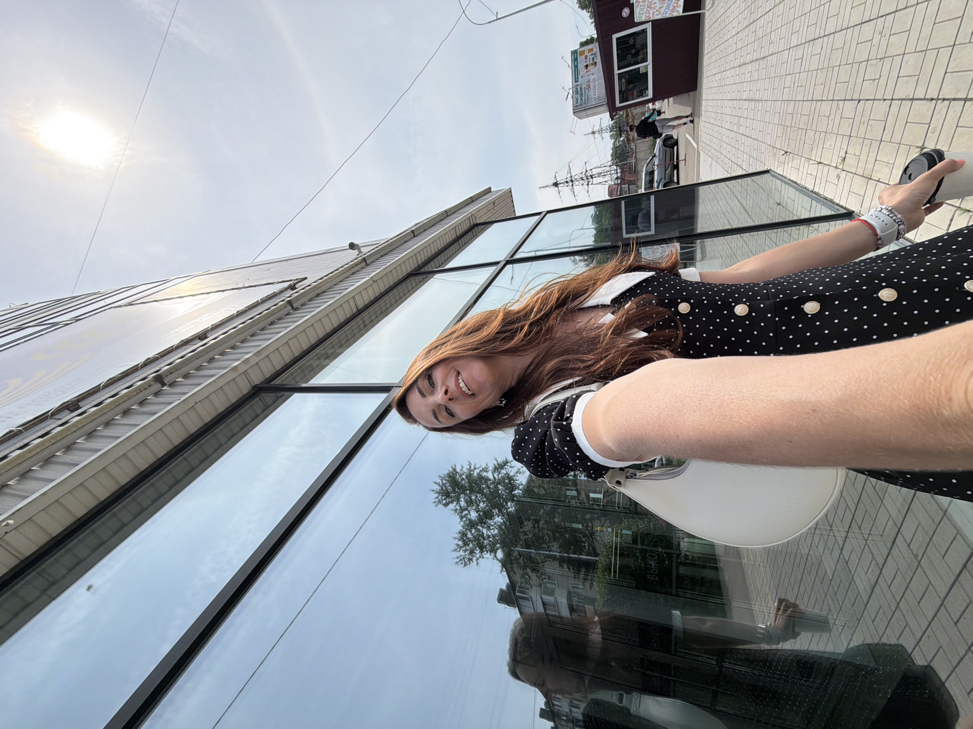 A woman with long, auburn hair takes a selfie outdoors, holding a white coffee cup in her right hand. She is wearing a black dress with white polka dots, a white collar, and gold buttons. She carries a white purse on her left shoulder. The woman is standing on a paved sidewalk in front of a building with large, reflective glass windows. In the reflection of the windows, a building with multiple stories and trees can be seen. The sky is bright and sunny, with some clouds visible. In the background, there are power lines, a car, and a small kiosk or booth. The location is identified as Birobidzhan, Russia.