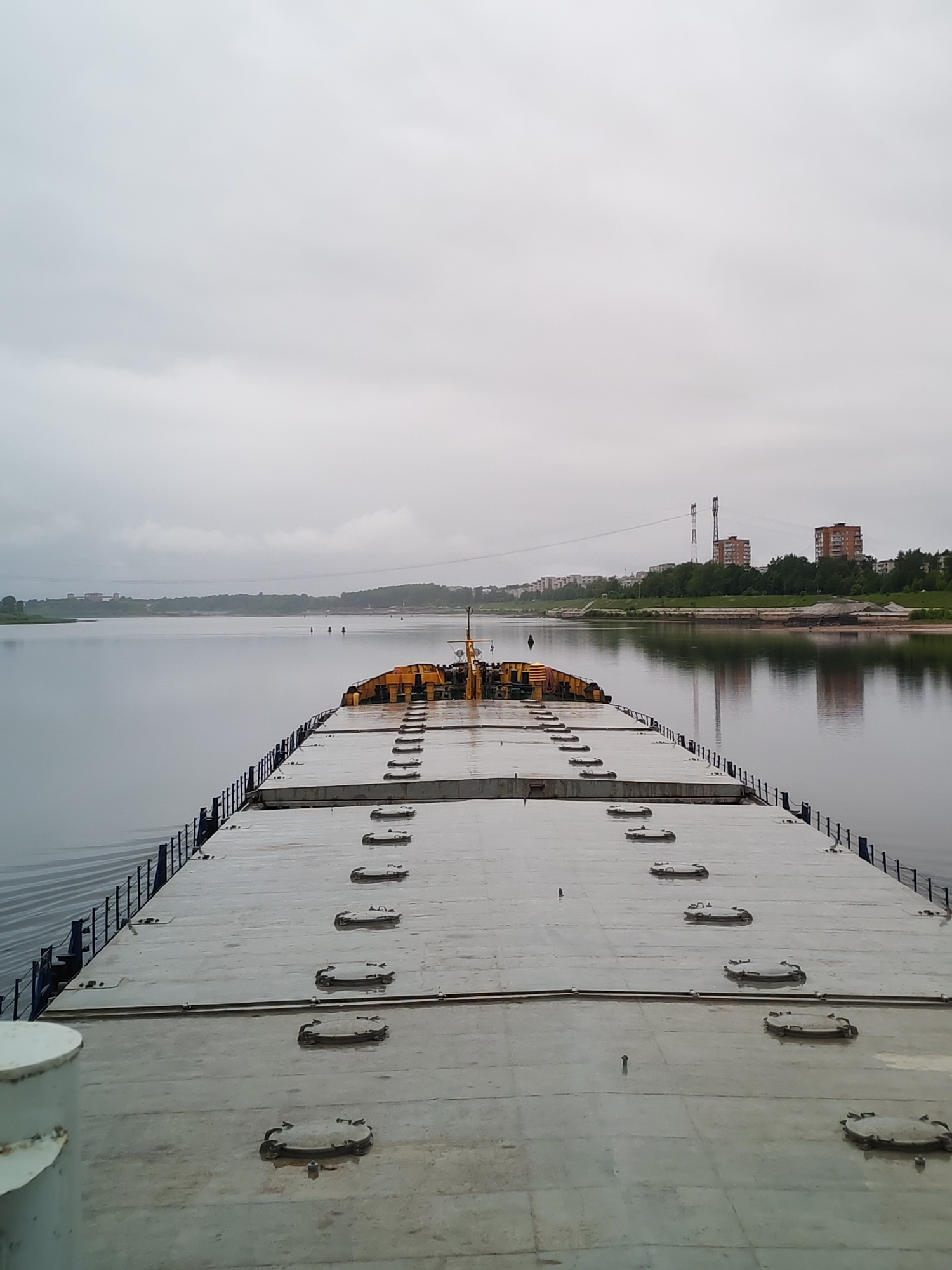The image depicts the flat deck of a barge on a body of water, likely a river or a large lake. The deck is made of large, rectangular concrete panels with a repeating pattern of circular, metal access hatches. A metal railing runs along the perimeter of the deck. In the distance, there are buildings and structures on the shoreline, which suggest an urban or developed area. The city of Bol'shie Mkhi, Russia is visible in the background, including specific structures such as tall smokestacks. The sky is overcast, and the water surface is calm with slight ripples. No people are visible in the foreground. The overall atmosphere appears to be somewhat somber due to the gray sky and the industrial setting.