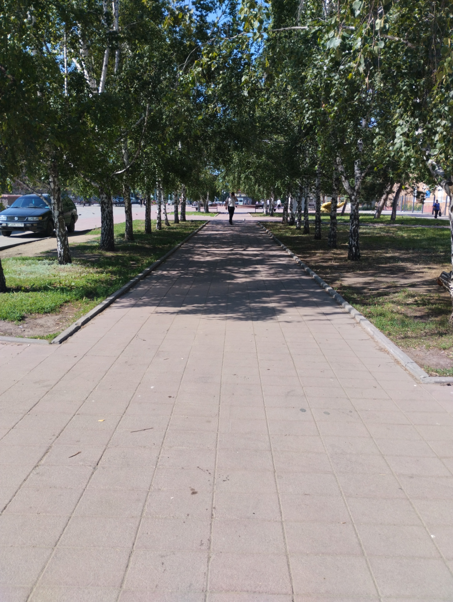 This is a full-shot, eye-level, outdoor photograph taken on a sunny day. The image depicts a paved walkway lined with birch trees on both sides. The walkway is made of square concrete tiles and stretches into the distance, forming an avenue. In the foreground, the tiles are clear, but further down the path, they are dappled with shadows cast by the trees.
On the left side of the walkway, a dark-colored sedan is parked. A few people are visible walking along the path in the middle ground. On the right side, there is a grassy area and more trees, with a glimpse of a fence and buildings in the background.
The scene appears to be a public park or a tree-lined street in the city of Kostanay, Kazakhstan, likely a place for recreation and pedestrian traffic. The overall atmosphere is serene and suggests a pleasant summer day.