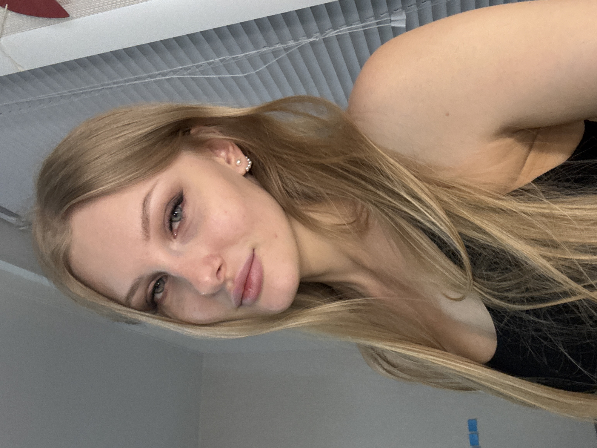 A blonde woman is shown in a close-up selfie, positioned against a backdrop of horizontal blinds. She is wearing a black top. The location is not identifiable as Perm, Russia.