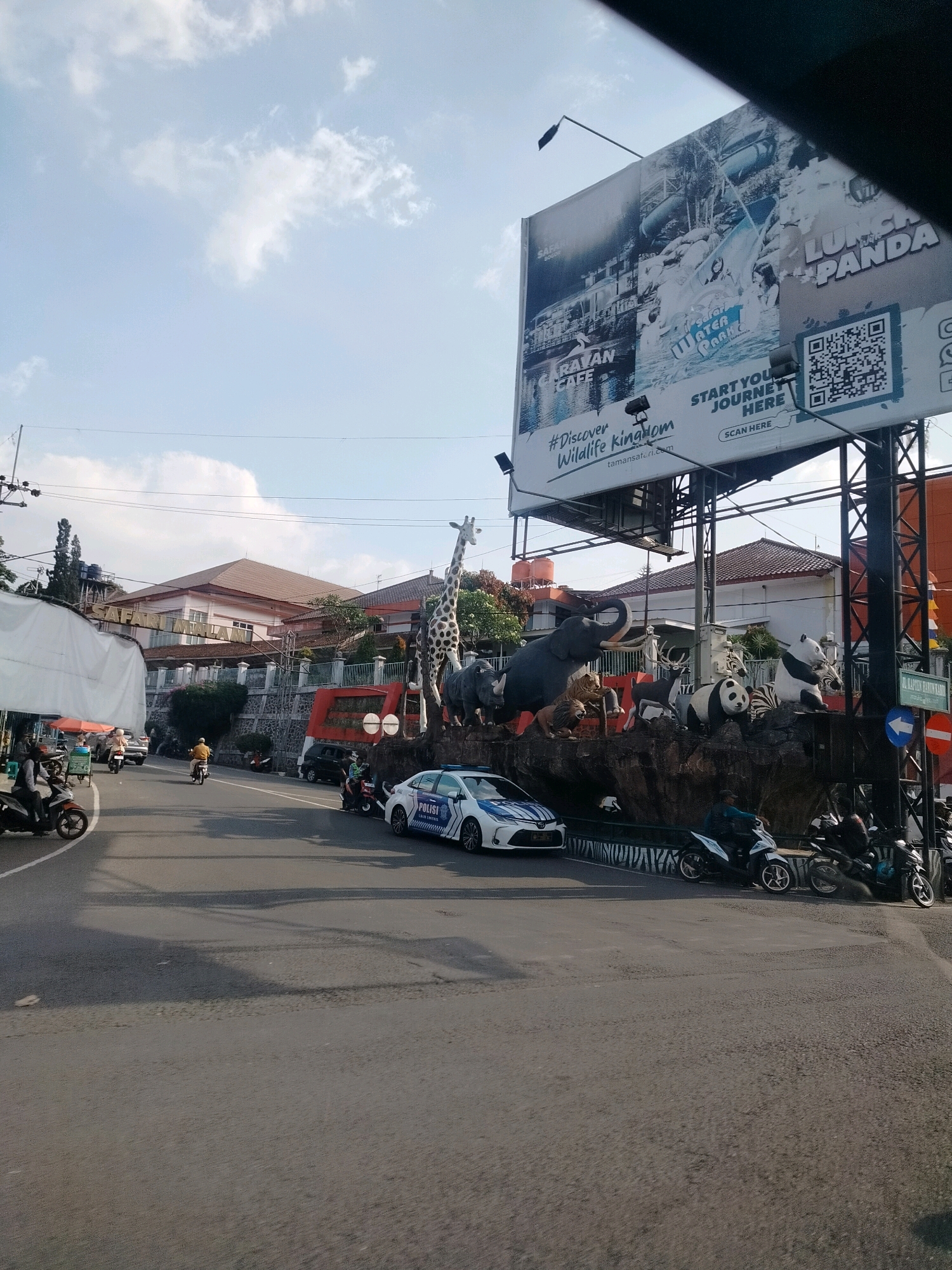 The image captures a street scene in Cisarua, Indonesia, on a sunny day with a partly cloudy sky. Dominating the upper right portion of the frame is a large billboard advertising "SAFARI MALAM" and "#Discover Wildlife Kingdom." The billboard features images of water slides, people enjoying water park activities, and mentions "PANDA" and "SAFARI WATER PARK." Below the billboard, a diorama-like display depicts various animal sculptures, including a rhino, lion, elephants, deer, zebras, and pandas, set against a rocky background.
In the foreground, a paved road shows traffic, with several motorcycles and a white police car with "POLISI" visible on its side. A person rides one motorcycle, and another motorcycle is parked with a rider. The police car is stationary in the right lane, with its front facing towards the viewer.
On the left side of the road, a white tent structure is partially visible, with people and carts underneath, suggesting a market or street vendor area. Buildings with tiled roofs are seen in the background, and utility poles with wires are present. The overall environment is urban, with elements of a tourist attraction prominently displayed.