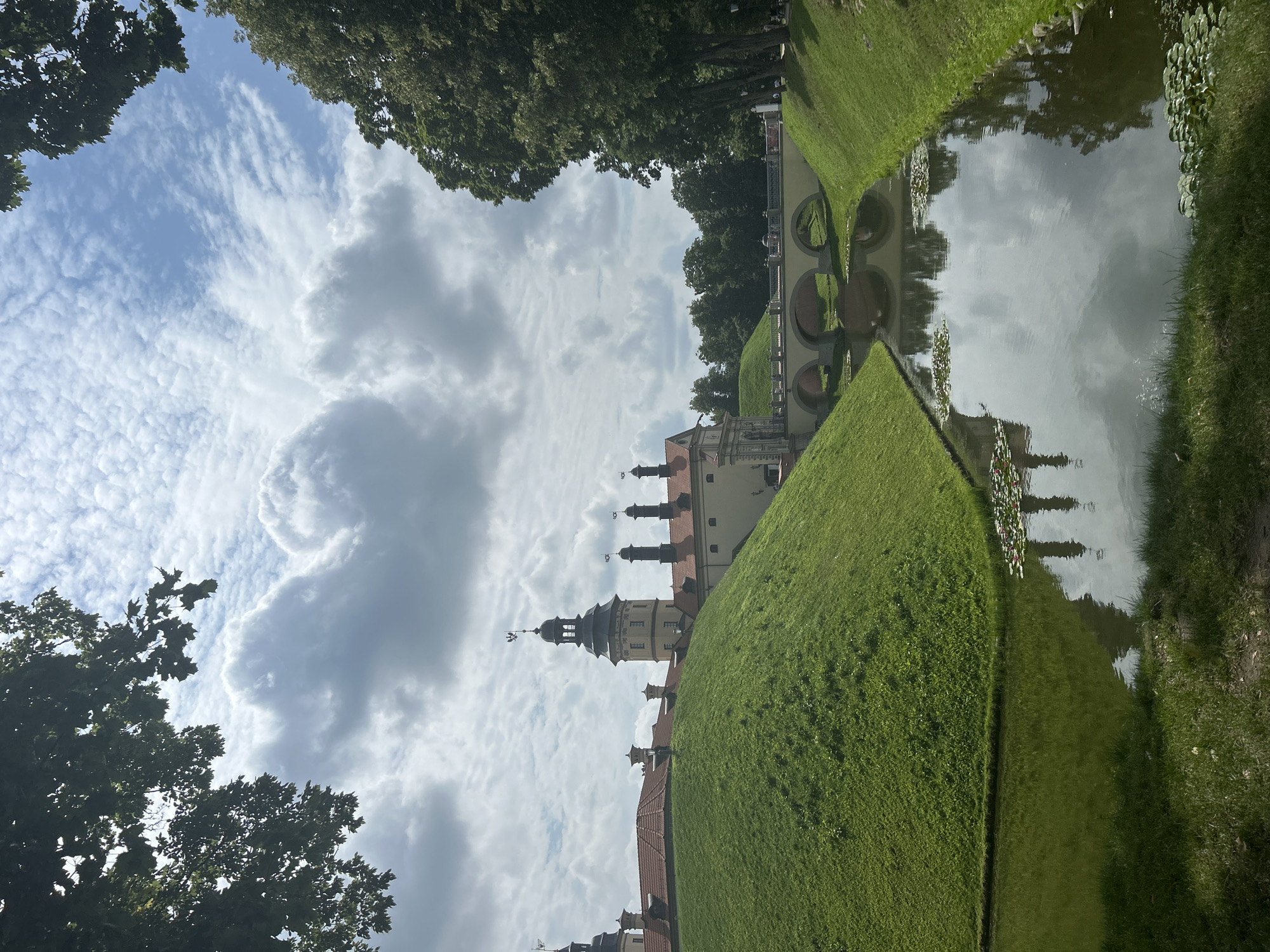 The image shows a view of Nesvizh Castle in Nesvizh, Belarus. The castle, with its red-tiled roofs and yellow walls, sits atop a large, grassy mound. A moat filled with still water, reflecting the sky and surrounding trees, surrounds the mound. A stone bridge with three arches spans the moat, connecting the castle grounds to the exterior. Lush green grass covers the slopes of the moat and the foreground, with some lily pads visible in the water. Trees frame the scene on the left and right, and a partly cloudy sky with dramatic cloud formations stretches across the top of the image. No people are prominently featured in the scene.