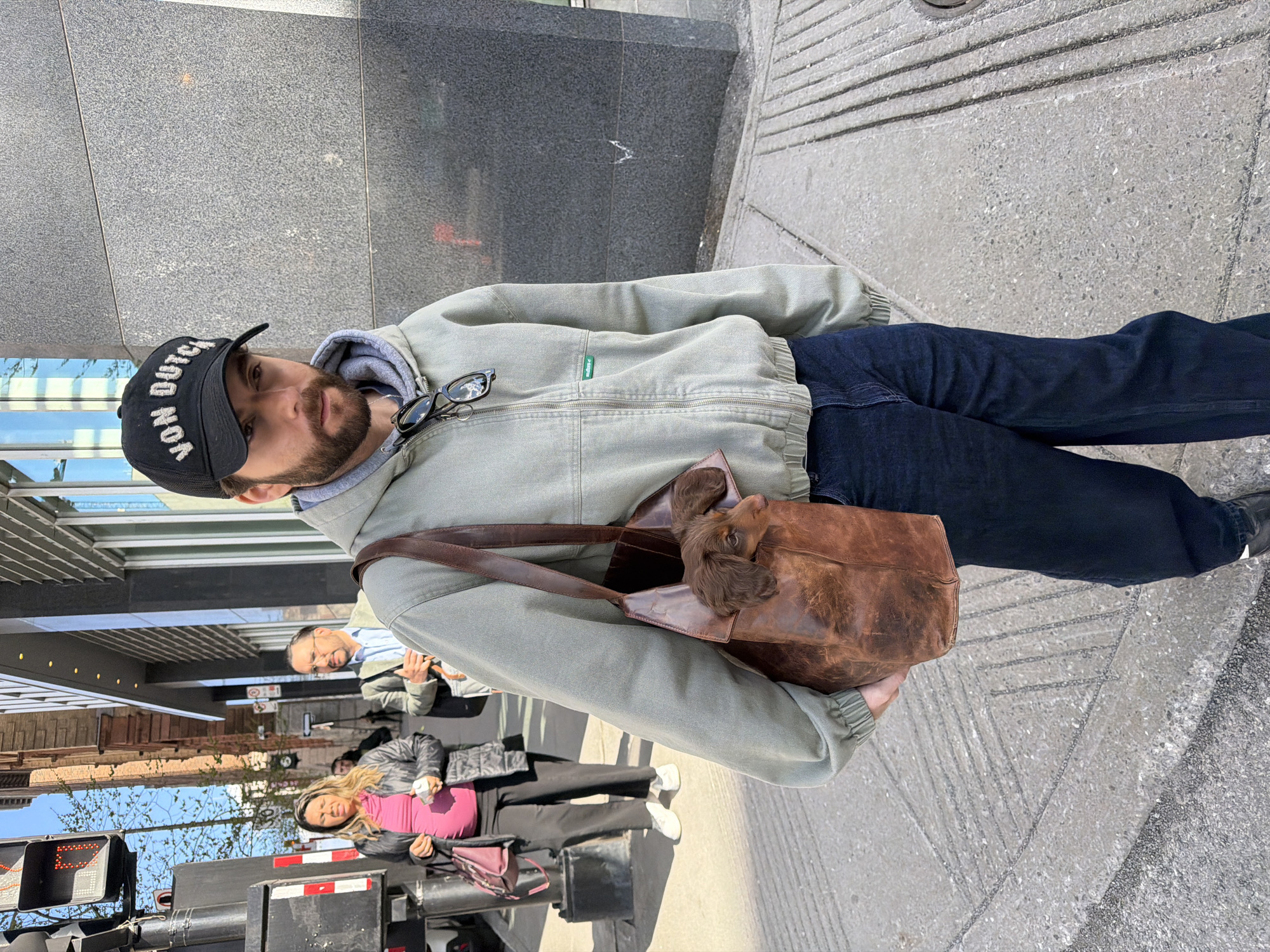 A person stands on a sidewalk near a building, wearing a black "Von Dutch" baseball cap, a light-colored jacket over a hooded sweatshirt, and dark jeans. They are carrying a brown leather bag, in which a brown dog is visible. In the background, there are other pedestrians on the sidewalk, a building facade, and a traffic light. Based on architectural style and street furniture, the location appears to be in Montreal, Canada.