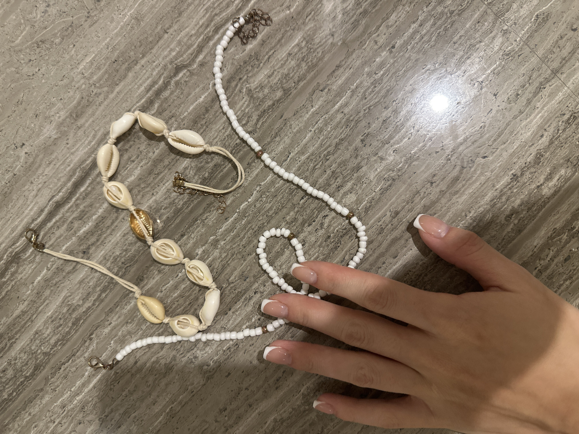 The image depicts two necklaces and a hand on a textured gray surface, likely a table or counter. One necklace consists of irregularly shaped, off-white shells strung together with beige cords and gold-tone clasps. The other necklace features small, round white beads interspersed with occasional small brown beads, and a silver-tone chain at one end. A hand with a French manicure is visible on the right side of the image, partially covering the beaded necklace. The location in the city of Unknown, Unknown is not discernible from the image.
