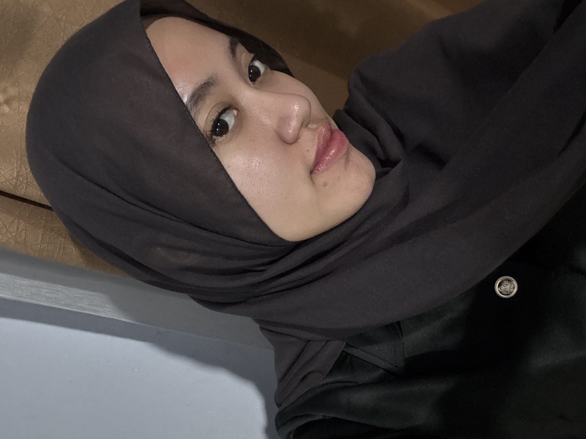 A young woman wearing a dark gray hijab and a black jacket takes a selfie. Her face is partially visible, with her eyes looking towards the camera. She is indoors, with a textured brown curtain in the background and a light gray wall with a shadow on the left side of the image. The scene appears to be a personal space in a residence in Gadingrejo, Indonesia.