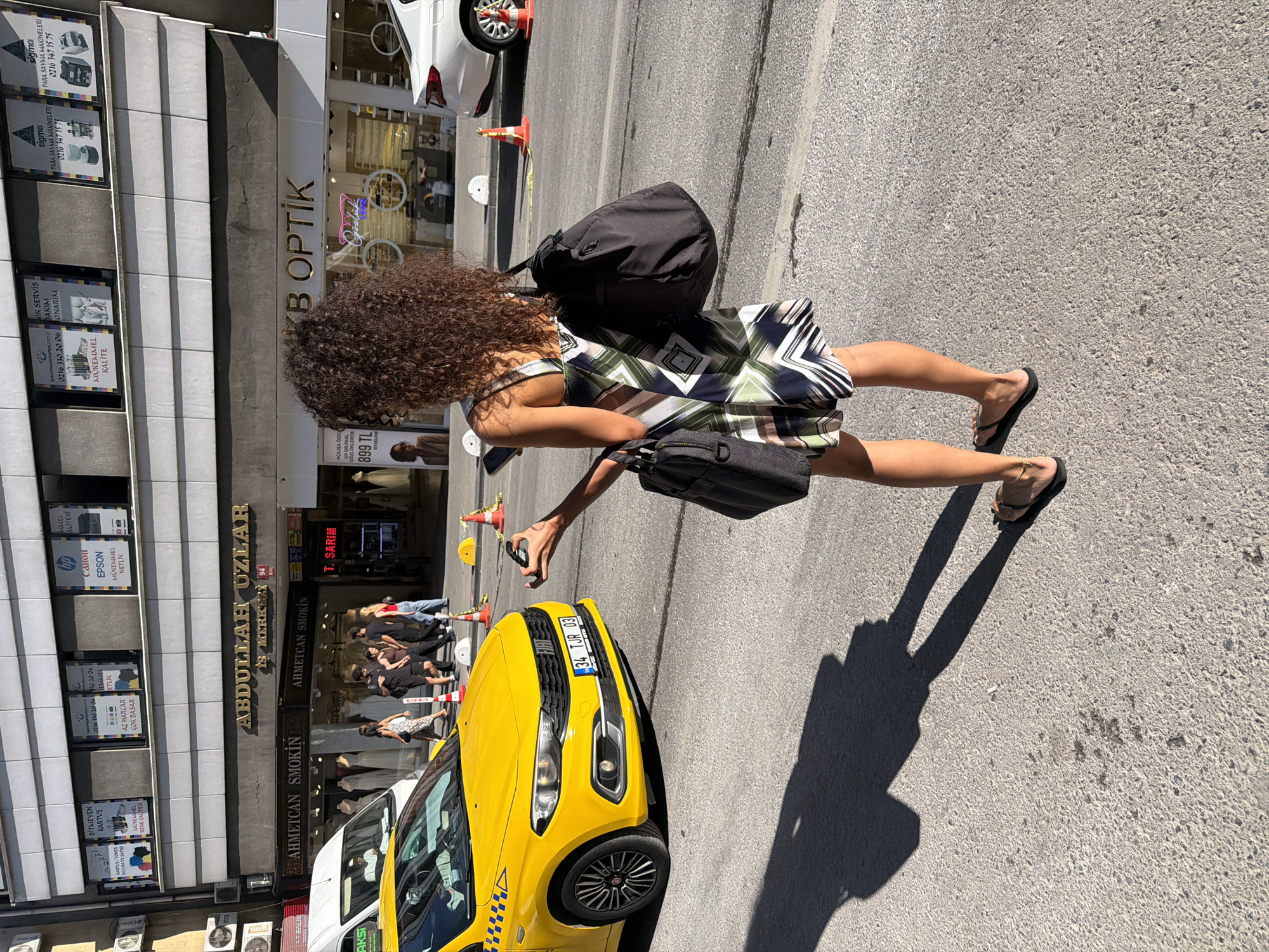 A woman with long, curly brown hair stands on a street in Kadıköy, Turkey, holding a phone and keys. She is wearing a patterned summer dress and flip-flops. She carries two bags: one over her left shoulder and a larger black bag over her right shoulder. In the foreground, her shadow is cast on the asphalt. To her left, a yellow taxi is parked. Behind her, buildings with shops and businesses line the street. One sign reads "ABDULLAH UZLAR" and another reads "B OPTIK". Other visible businesses include "AHMETCAN SMOKIN" and "T. SARIM". Several people are walking in the background. Orange traffic cones are visible on the street.