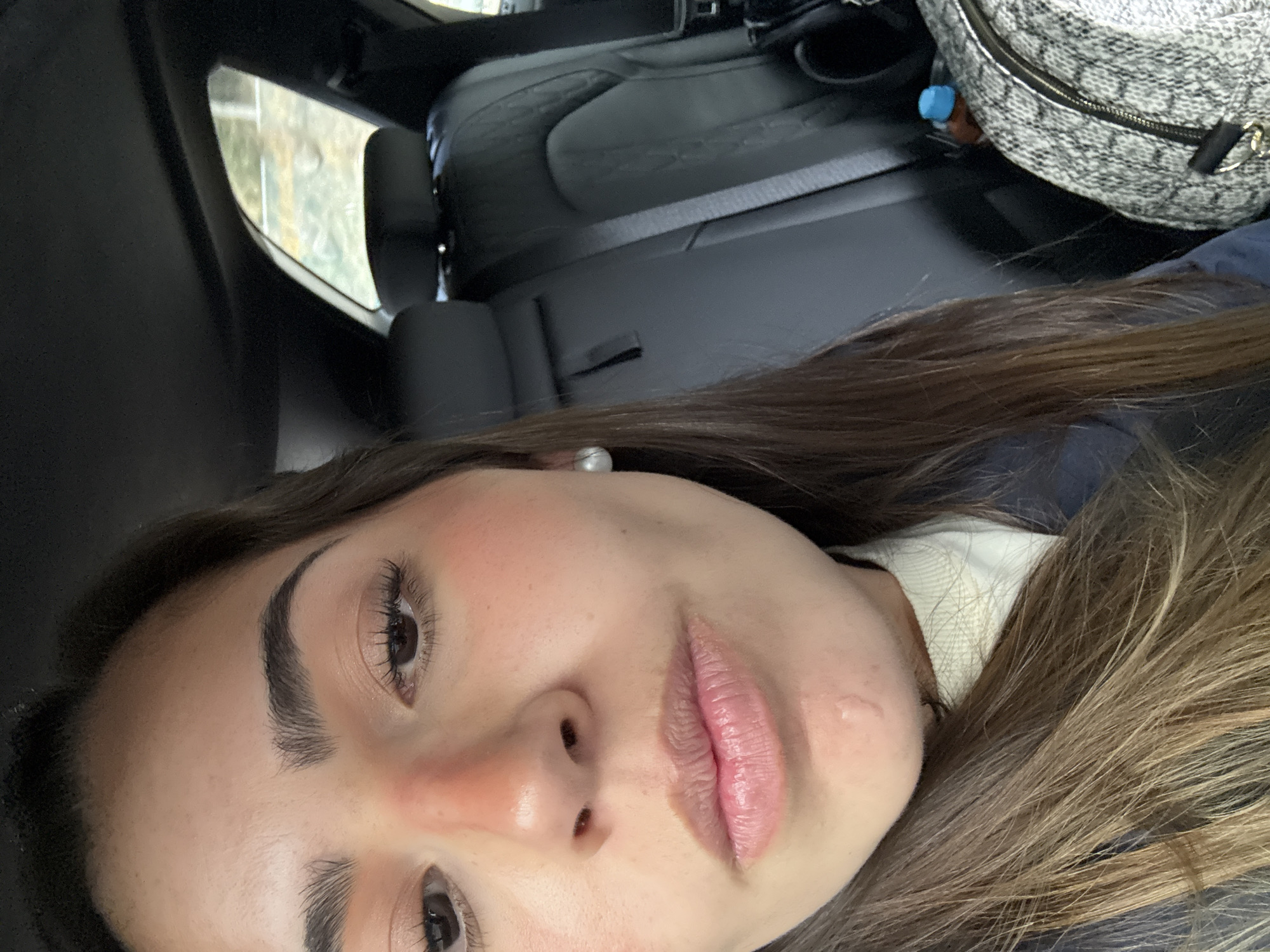 A close-up, tilted portrait of a woman with brown hair, fair skin, and dark eyes inside a vehicle. She is facing the camera. She is wearing a white top underneath a dark colored jacket. The interior of the car consists of black leather seats. A patterned bag is visible in the background. The location of the image is not discernible from the scene and therefore cannot be determined to be in San Marcos Huixtoco, Mexico.