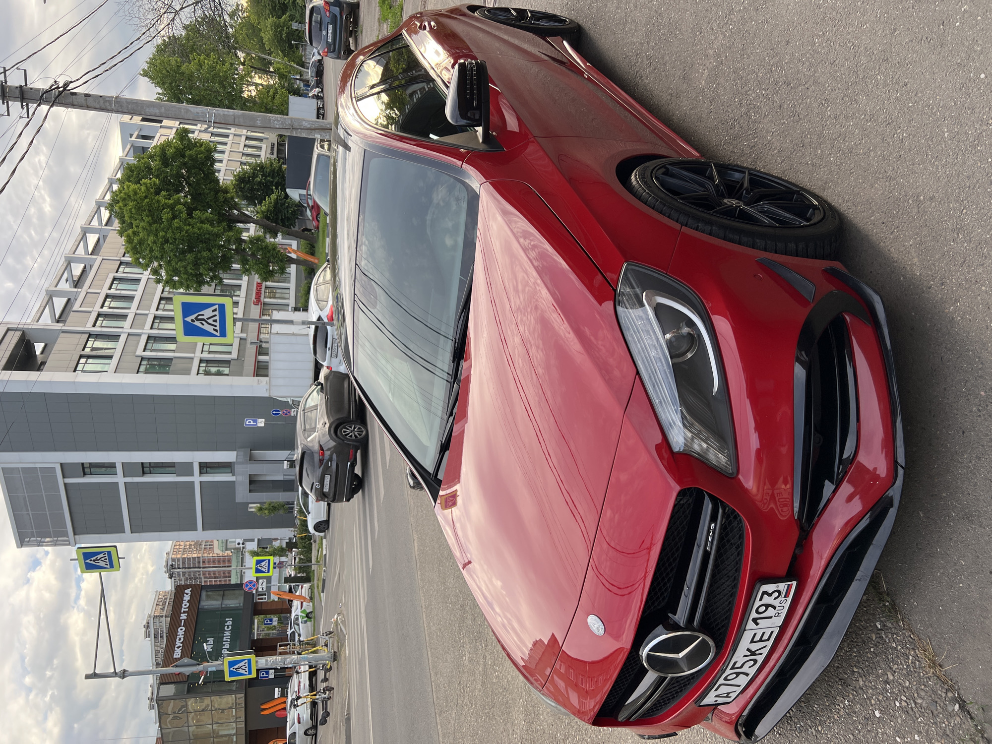 The image, taken in a portrait orientation, captures a vibrant red Mercedes-Benz car, possibly an AMG model, parked on the side of an urban street. The car is the primary subject, featuring black multi-spoke alloy wheels and a sporty front bumper with black aerodynamic elements. Its Russian license plate, "A795KE 193 RUS," is clearly visible on the front. The "AMG" badge is also visible on the grille.
The scene depicts a typical city environment during daytime, under an overcast or partly cloudy sky. Modern multi-story buildings, varying in architectural style with large windows and grey/white facades, line the street. Trees and green foliage are interspersed along the sidewalks. A prominent street pole with power lines runs across the upper left portion of the image.
Other vehicles are visible in the background, including parked cars and several electric scooters or bicycles parked on the sidewalk. Numerous road signs are present, including multiple blue-and-white pedestrian crossing signs (a white walking person within a triangle on a blue square) and blue parking signs marked with a white 'P'.
Visible Cyrillic text on storefronts includes "ВКУСНО - И ТОЧКА" (VKUSNO - I TOCHKA), identifying a well-known Russian fast-food chain. Another partial sign reads "МЫ" (MY) and "КРЫЛИСЬ!" (KRYLIS!), likely part of a "WE ARE OPEN!" message. No people are discernible in the frame. The overall setting conveys a common street scene in Krasnodar, Russia.