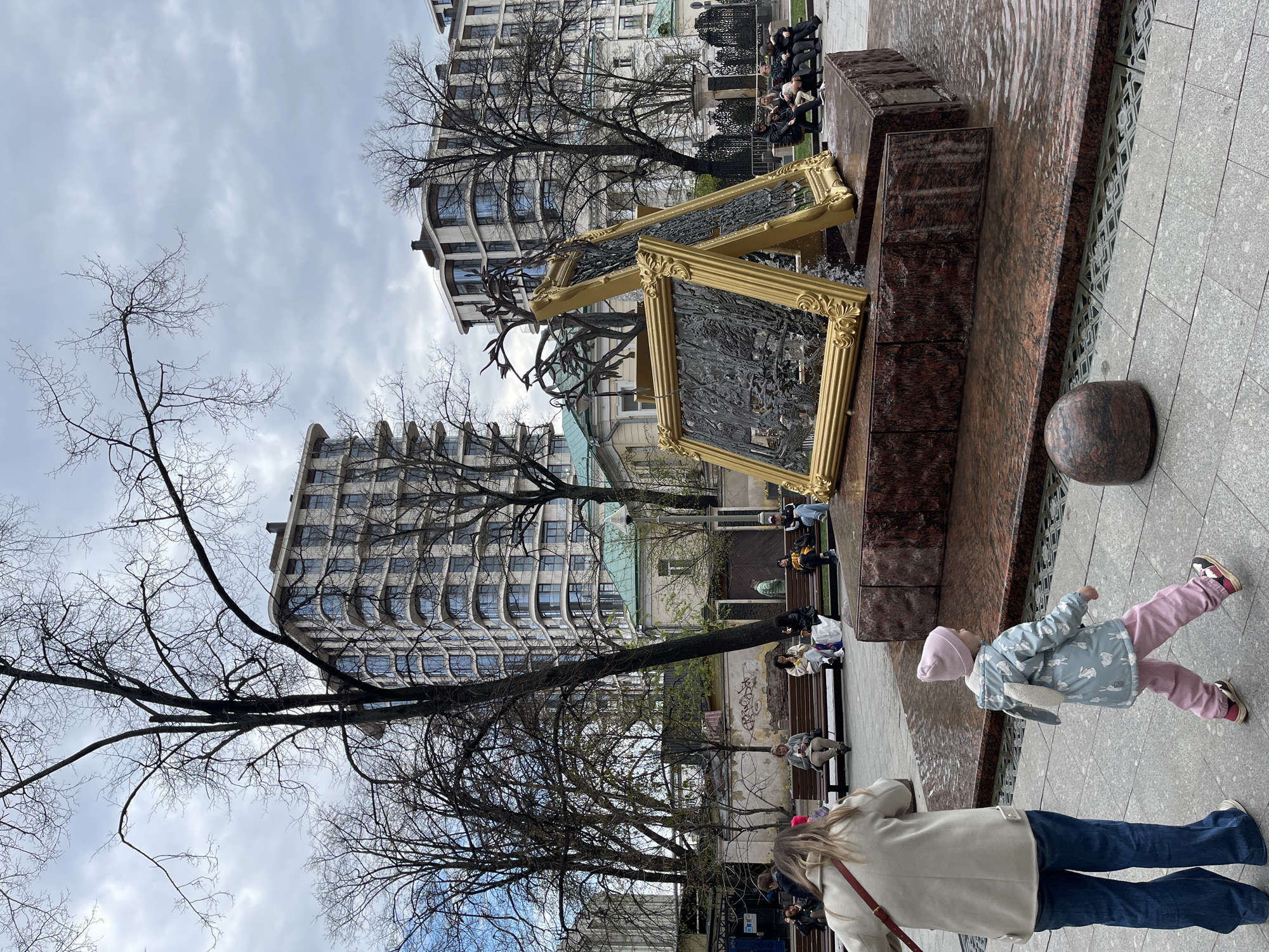 The image captures a scene in Moscow, Russia, featuring a unique water fountain adorned with golden picture frames containing bas-relief sculptures. The fountain is constructed from dark red granite blocks with water flowing over the sides. In the background, there are several modern buildings with curved architecture, interspersed with bare trees characteristic of early spring or late autumn.
Several people are visible, including a woman with long blonde hair wearing a light-colored coat and blue jeans, with a red purse or bag. A child in pink pants and a light blue jacket with bunny designs is walking on the sidewalk, while other individuals are sitting on benches near the fountain. The sky is overcast with heavy clouds. The scene appears to be a public space or park where people are relaxing and enjoying the urban environment.