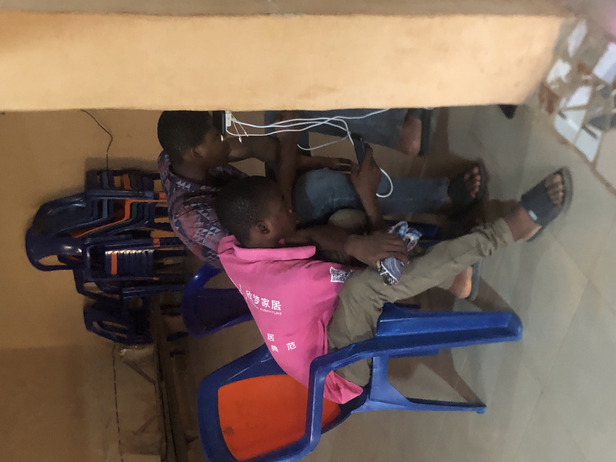 Two people are sitting under a table in Nkwelle Ezunaka, Nigeria. One person wears a pink shirt with Chinese characters and olive pants and sits in a blue and orange plastic chair, their feet resting on another chair. The second person wears a patterned shirt and jeans. A stack of blue and orange chairs is visible behind them. There are wires and electronic devices under the table. The floor is light-colored tile.