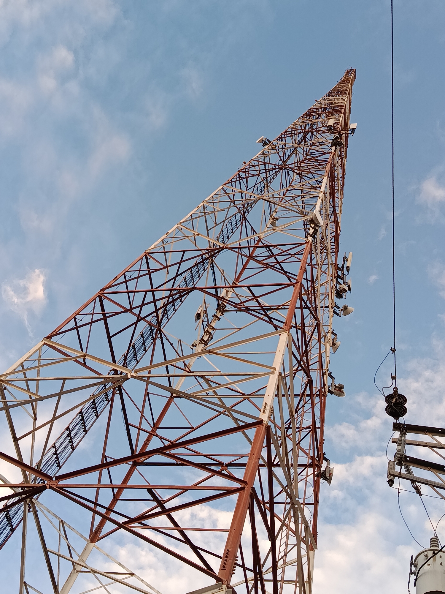A towering lattice cell tower, painted in alternating sections of red and white, dominates the frame, reaching towards a pale blue sky dotted with wispy white clouds. The perspective is from below, looking up at the immense structure, emphasizing its height and intricate metal framework. Several antennas and communication equipment are mounted at various levels on the tower. To the right of the cell tower, a thick black cable runs vertically, disappearing out of frame. Further to the right and slightly lower, a portion of an electrical utility pole with insulators and a transformer is visible. The scene appears to be an outdoor industrial or telecommunications area, possibly on the outskirts or within the city of Juan Griego, Venezuela, given the context. No people are directly visible in the frame, and no specific actions or interactions are depicted.