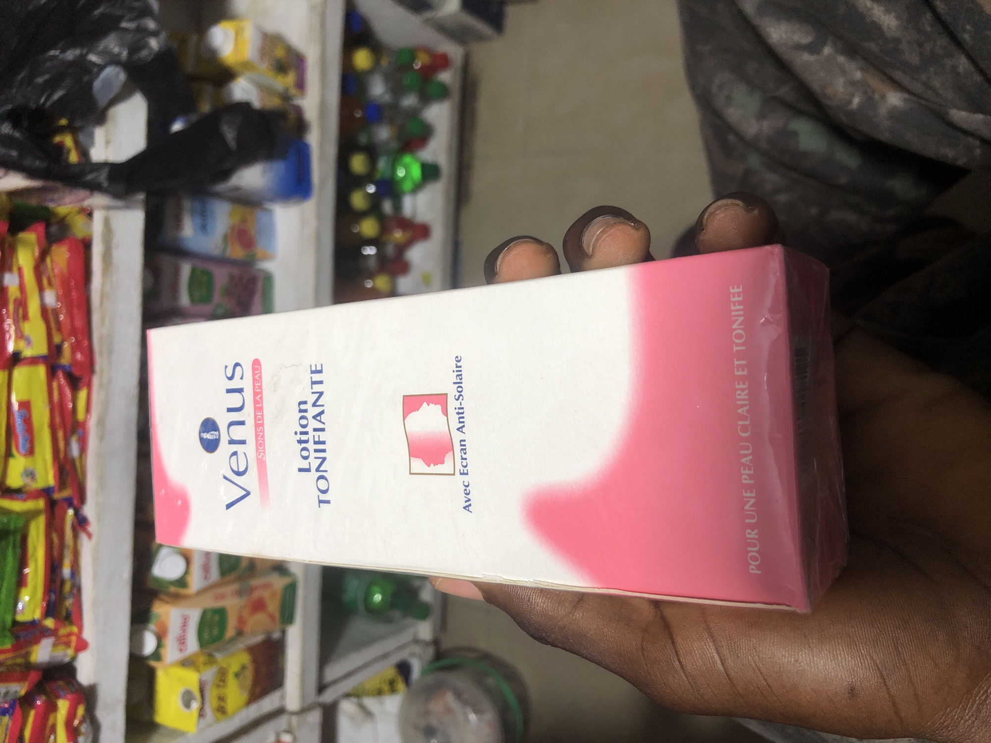 The image shows a person holding a box of "Venus Lotion Tonifiante" in what appears to be a retail setting. The box is pink and white with French text. In the background, shelves are stocked with various products, including packaged snacks and beverages, suggesting a small shop or market stall, possibly in Ado, Nigeria. The lighting is somewhat dim, and the focus is on the product in the person's hand.