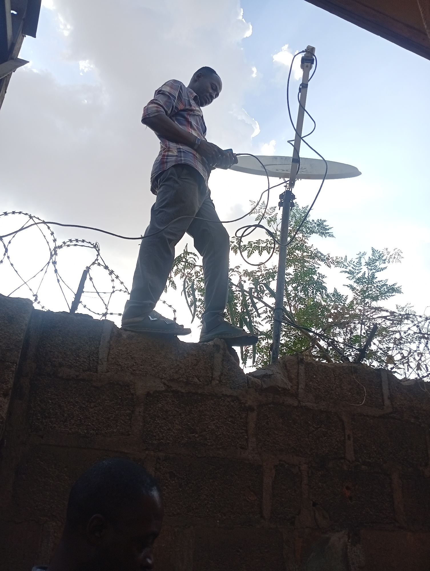 A man in a plaid shirt and gray pants stands on a brick wall topped with barbed wire, adjusting a satellite dish on a pole. A partial view of another person’s head appears in the foreground. The background features a cloudy sky and sparse foliage. The location is Sabon Tasha, Nigeria.