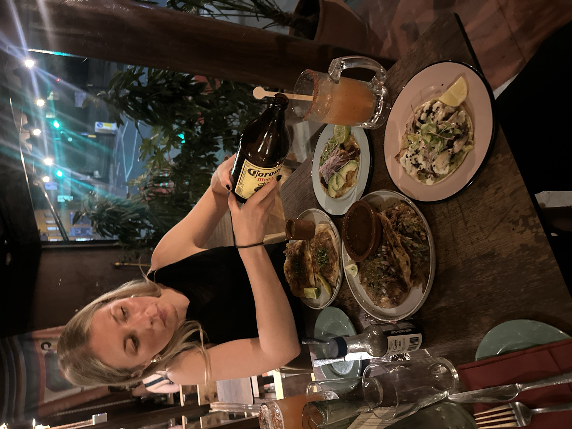 Here is a detailed description of the image:
The image captures a woman holding a Corona beer bottle over a table laden with food and drinks. The table is primarily wooden and features a variety of plates with what appears to be tacos, a beer mug filled with a yellow liquid, and a bottle of hot sauce. The woman is fair-skinned with blonde hair and is wearing a black sleeveless top. She is looking at the beer bottle in her hands.
The table also has glasses, cutlery, and other dining accessories. In the background, a window provides a glimpse of a cityscape at night, showing lights and what appear to be buildings, giving the impression that the scene is inside a restaurant or bar.
Although no specific location can be determined from the image, the scene may represent a restaurant in any major city, including London, United Kingdom.