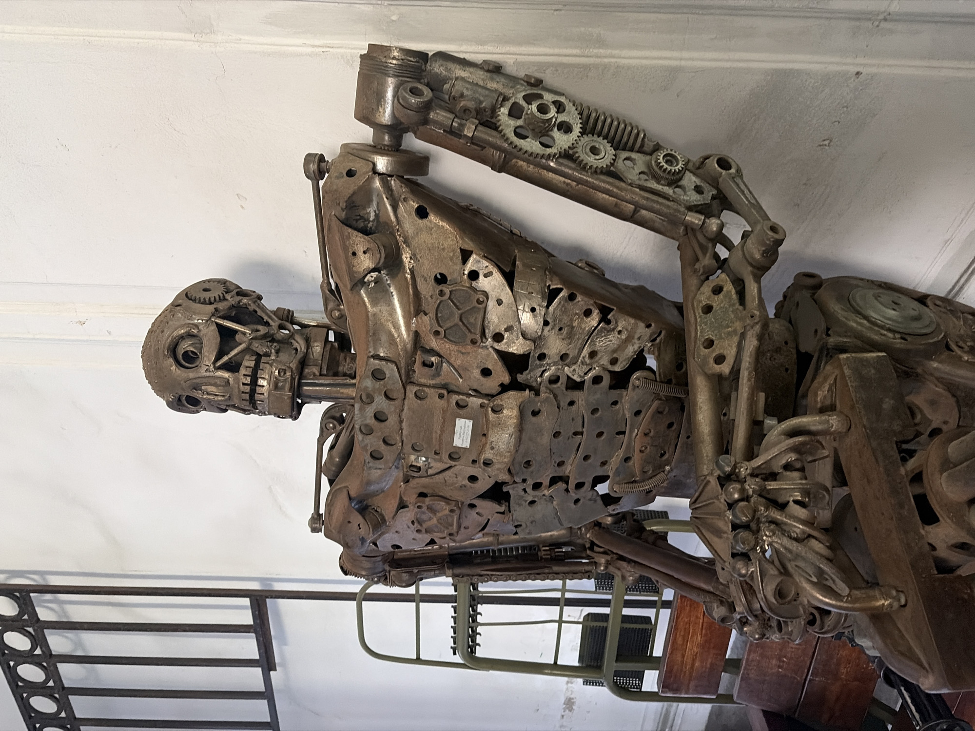 The image shows a sculpture of a skeletal robot made of metal parts and gears, positioned indoors against a white wall. It's affixed to a display apparatus and mounted on a brown wooden platform, which is integrated into a green, ladder-like frame. The sculpture appears to be in Saint Petersburg, Russia.