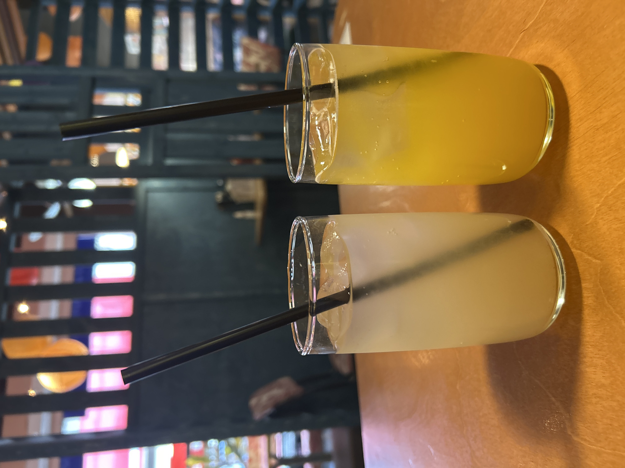 Two clear glasses filled with liquid are resting on a wooden surface. The top glass contains a yellow liquid, possibly orange juice or lemonade, while the lower glass holds a lighter, opaque liquid. Each glass has a black straw. The background reveals a dark, wooden structure with colorful, blurred lights or designs, suggesting an indoor setting. The image is taken at an angle and appears to be slightly tilted. It is unlikely to be from Izhevsk, Russia based on the objects and the setting.