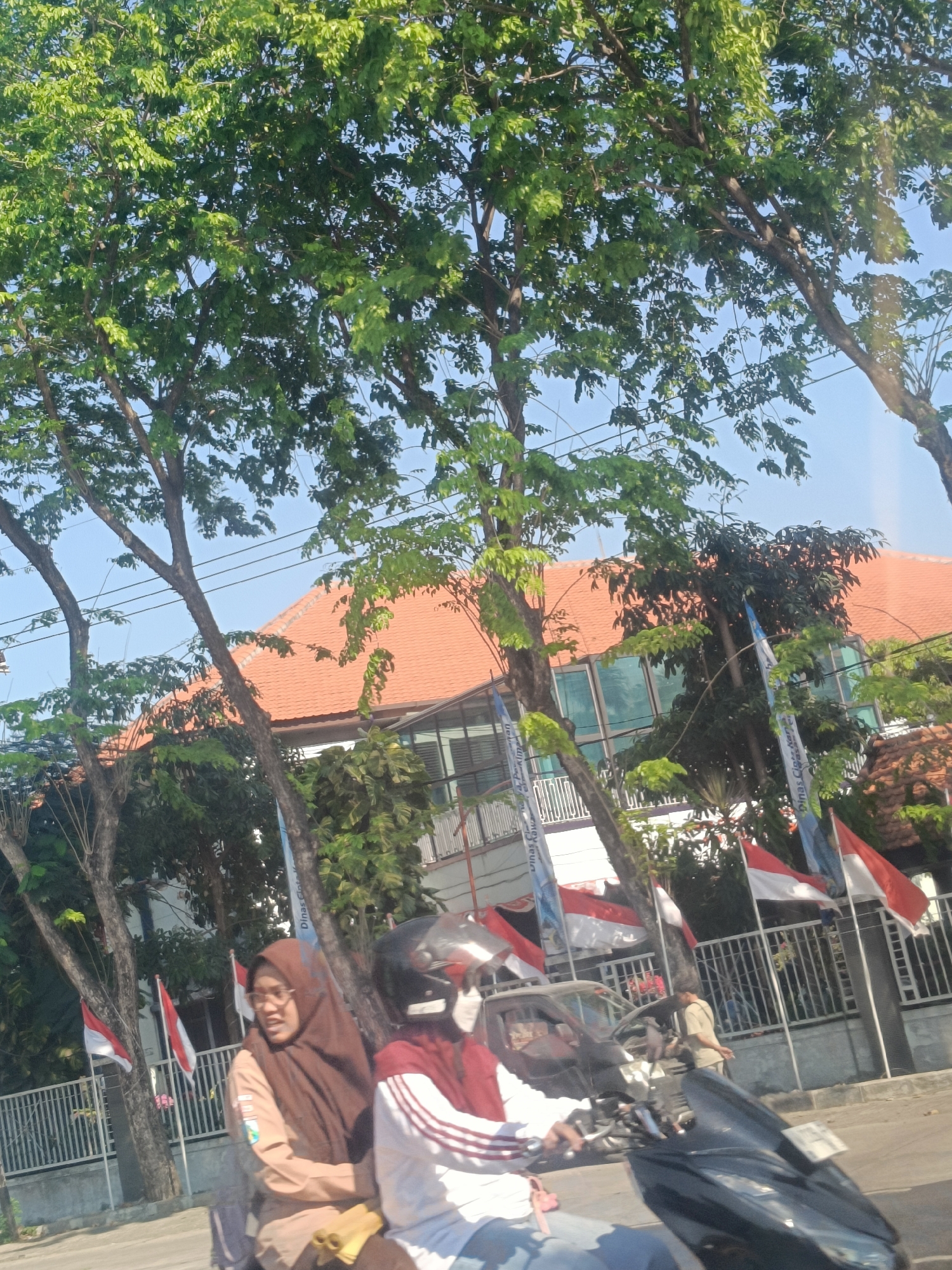 The image depicts a street scene in Dahanrejo, Indonesia. Two individuals are riding on a motorcycle. The person in the foreground, driving the motorcycle, is wearing a black helmet and a maroon and white striped shirt. The person behind them, a woman, is wearing a brown hijab and a tan uniform with a patch on the left sleeve, and glasses. They appear to be students. In the background, a building with an orange tiled roof and large glass windows is visible, partially obscured by trees. Several Indonesian flags (red and white) are displayed along a fence in front of the building. The trees have lush green foliage and the sky is clear blue. The overall scene suggests a typical day on a street in an Indonesian town.