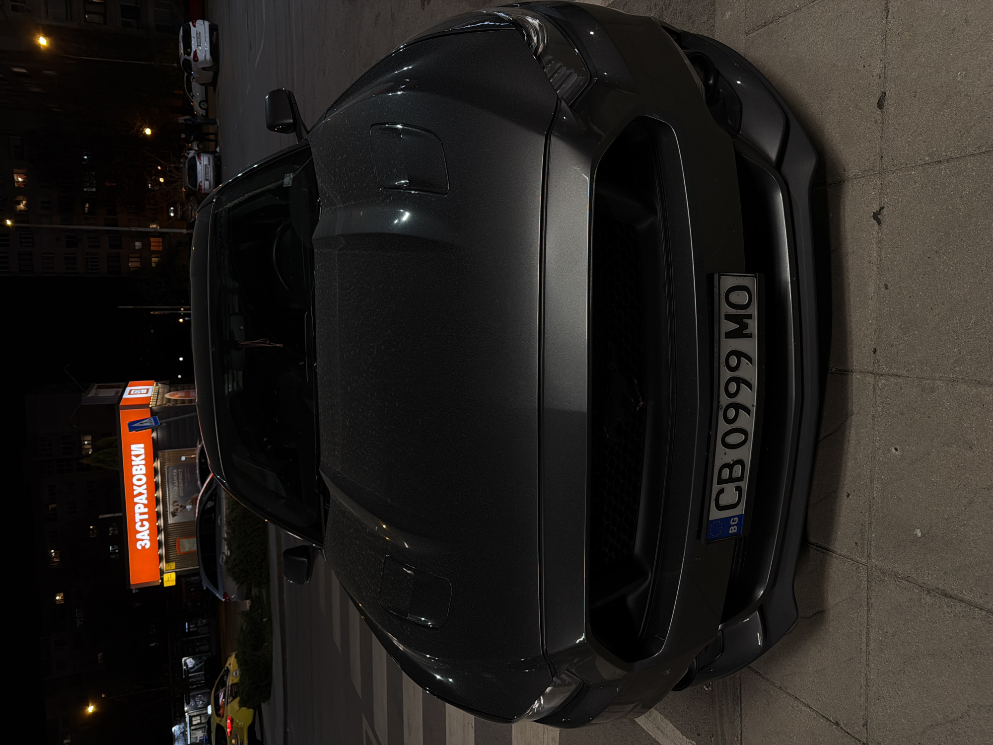 The image captures a dark-colored Ford Mustang, likely an S550 generation, parked at night in an urban setting. The car is positioned with its front fascia and hood prominent, appearing slightly wet or dusty with moisture. The overall lighting is dim, sourced from streetlights and illuminated buildings in the background.
A Bulgarian license plate, "CB 0999 MO," is clearly visible on the front bumper, with "BG" and the EU stars indicating its country of origin. In the background, other vehicles are discernible, including a white SUV and a yellow taxi, suggesting a busy street or parking area. Residential or commercial buildings with glowing windows line the street, contributing to the ambient light.
A prominent, large orange sign with red Cyrillic text reads "ЗАСТРАХОВКИ" (ZASTRAKHOVKI), which translates to "INSURANCE" in Bulgarian. This sign is a key indicator of the location being in Sofia, Bulgaria, aligning with the "CB" district code on the license plate. The time of day is clearly night, characterized by the dark sky and reliance on artificial light sources. The scene is otherwise still, with no apparent active events beyond the parked vehicles. The pavement beneath the car is visible, a standard concrete block design.