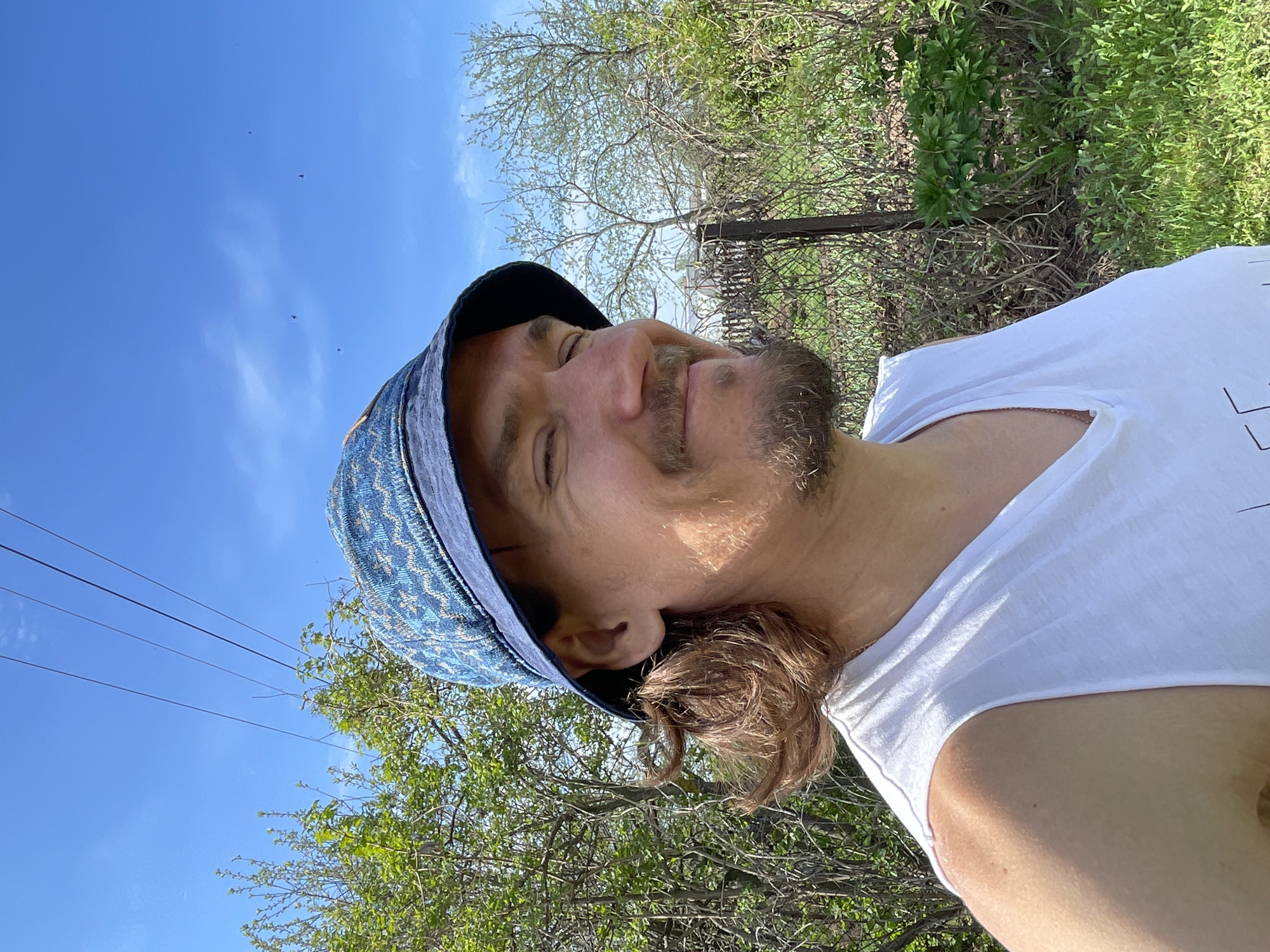 A medium outdoor shot, rotated counterclockwise, features a light-skinned individual with a mustache and goatee wearing a blue patterned bucket hat and a white t-shirt. The individual’s eyes are closed and the head is tilted back. Behind the person, the lower portion of the frame shows green grass and trees with sparse green foliage. The upper part of the background is a clear blue sky with thin power lines. The location is Перевозное, Russia, based on the presence of power lines and indigenous tree varieties.