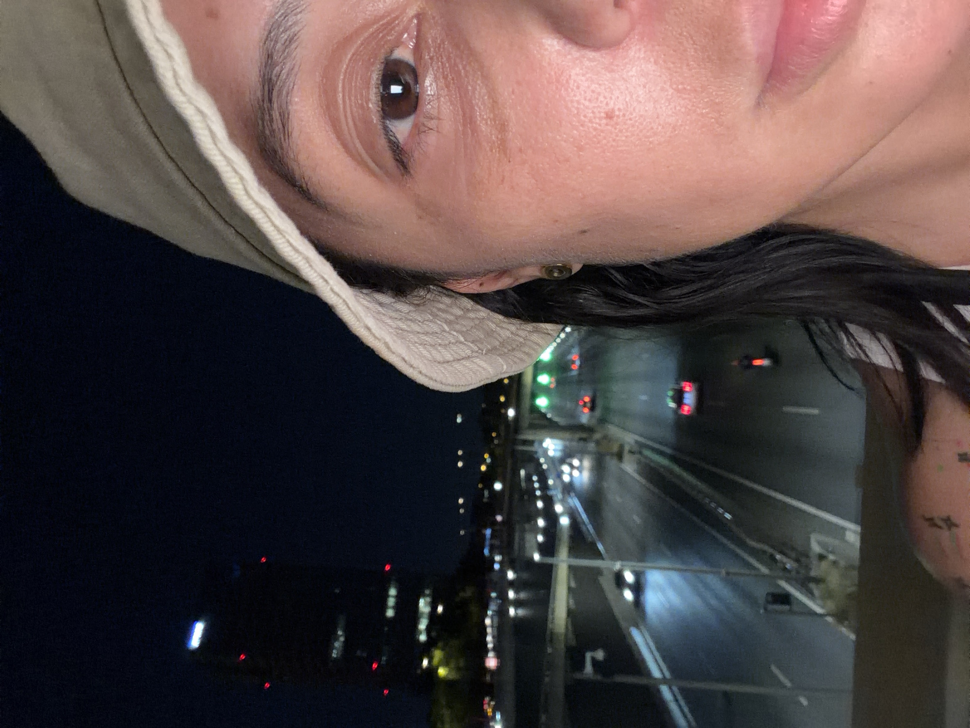 A close-up, angled photo of a person's face dominates the top of the image. The person wears a light-colored bucket hat. Below the person, a nighttime city scene is visible. A multilane highway stretches into the distance, with cars leaving streaks of light. Adjacent to the highway, buildings with lighted windows are seen, indicating an urban environment. Based on the information provided, this is in L'Hospitalet de Llobregat, Spain.