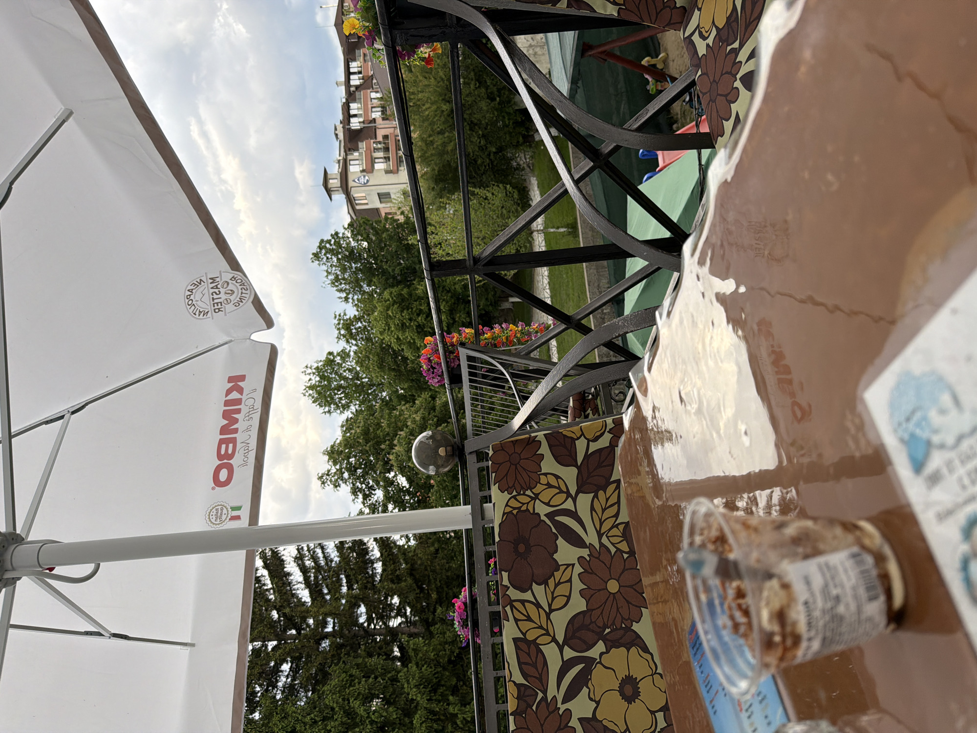 The image captures an outdoor cafe or restaurant setting, looking out from a terrace or balcony under a bright, cloudy daytime sky. The perspective is tilted, making the ground appear on the left and the sky on the right.
In the immediate foreground, a reflective brown tabletop holds a small, clear plastic cup containing a dessert or snack, and a partially visible menu or paper item with blue graphics. A patterned cushion or table runner, adorned with brown and yellow floral designs, is also on the table.
To the left, a large white outdoor umbrella provides shade. The brand "KIMBO" with "Il caffè di Napoli" is clearly visible in red text on one panel. Another circular logo, displaying "MASTER BARISTA" and "QUALITA SUPERIORE" (partially obscured and reversed), is on another section of the umbrella.
Beyond the table, a decorative black wrought iron railing, embellished with flower boxes overflowing with colorful petunias, frames the view. A spherical light fixture is attached to the railing. Below, a lush green landscape stretches out, featuring various trees, including tall pines, and a grassy area. In the distance, several buildings with balconies can be seen. One building stands out with a distinctive rounded tower or roof feature. Partially visible behind the railing and among the trees is a children's play area with red, blue, and green elements.
The scene evokes a peaceful and relaxing ambiance, characteristic of enjoying a break outdoors in pleasant weather.