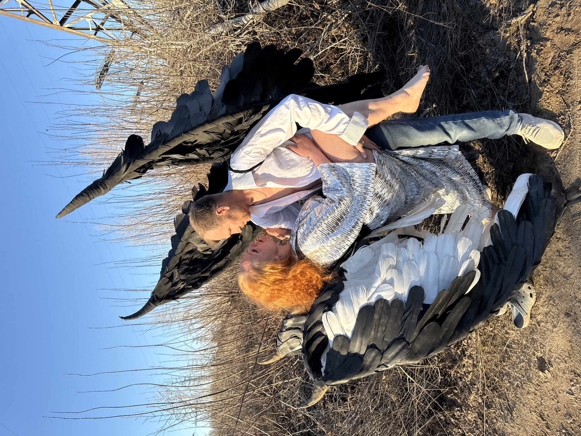 The image shows two individuals embracing on the ground outdoors. One individual, with red hair, wears a silver sequined shirt and black and white wings. The other individual, with blonde hair, wears a white shirt and jeans and has one bare foot. They are lying on dry ground covered with dead twigs and leaves. One individual has black wings behind them. A metal power line structure is visible in the background, extending into the blue sky. A beige Crocs-style shoe is also on the ground nearby. The photograph was taken in the city of Chaikovsky, Russia.