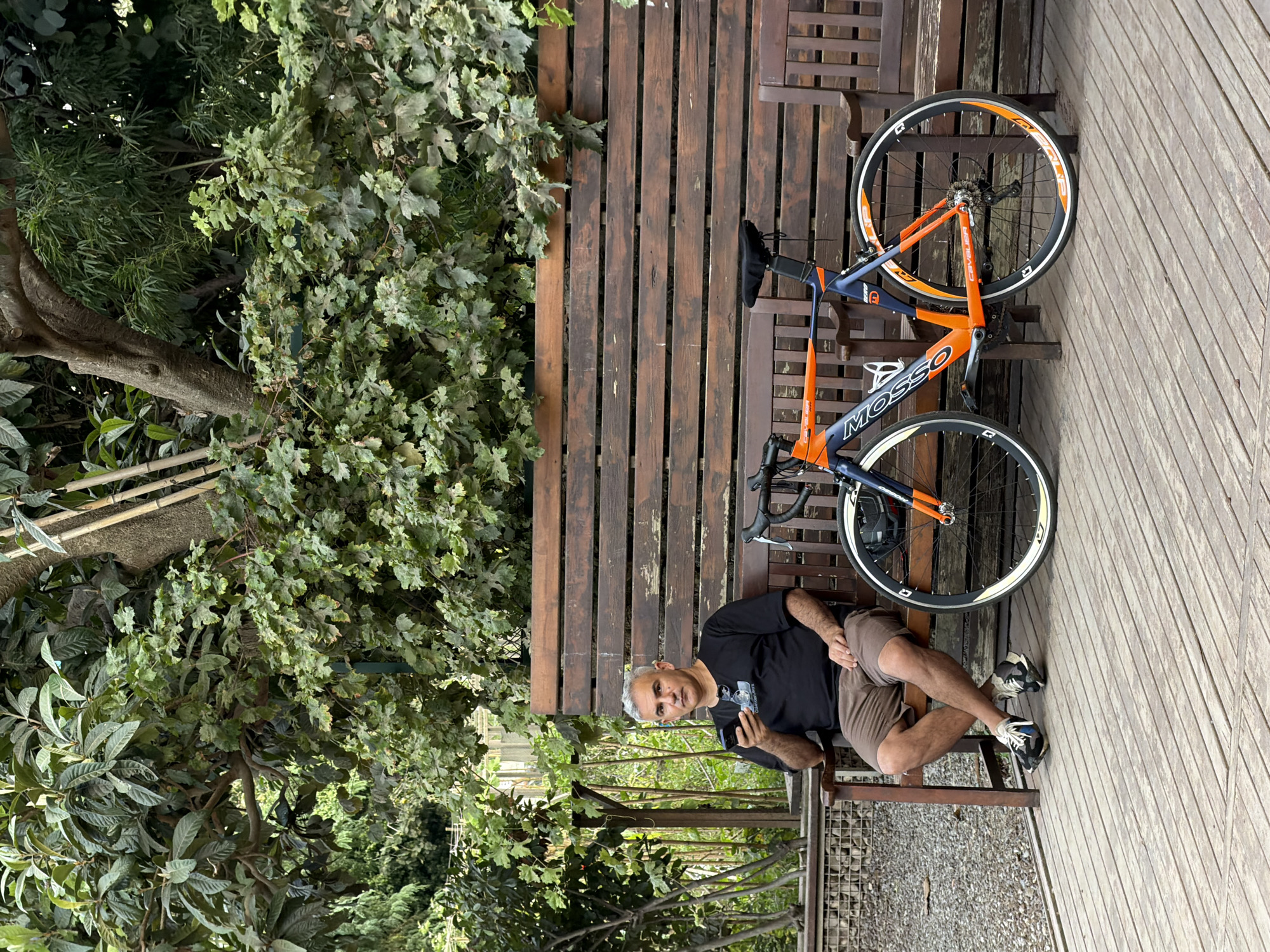 A man with graying hair is seated on a wooden bench, holding a smartphone. He is wearing a black t-shirt and brown shorts. A bicycle with an orange and blue frame is parked next to him. The scene appears to be outdoors, with lush green foliage and a wooden deck. The overall setting suggests a park or a quiet outdoor recreational area.