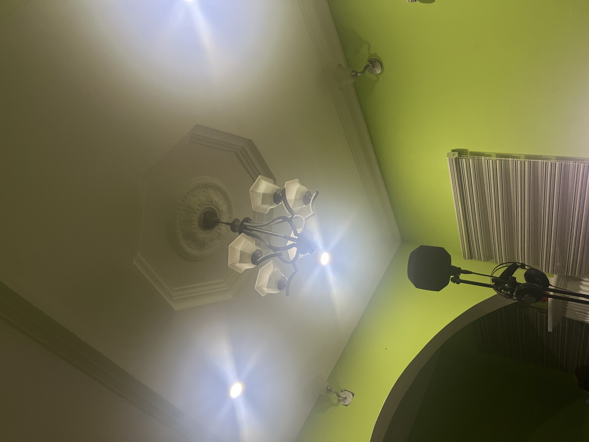 The image shows a view of a room's ceiling and wall. The ceiling is white with a decorative medallion and a chandelier with multiple lights. The wall is green with visible window blinds. A microphone and headset are attached to a stand near the wall. There is no indication of the location being in Abuja, Nigeria. There are no people visible in the image.