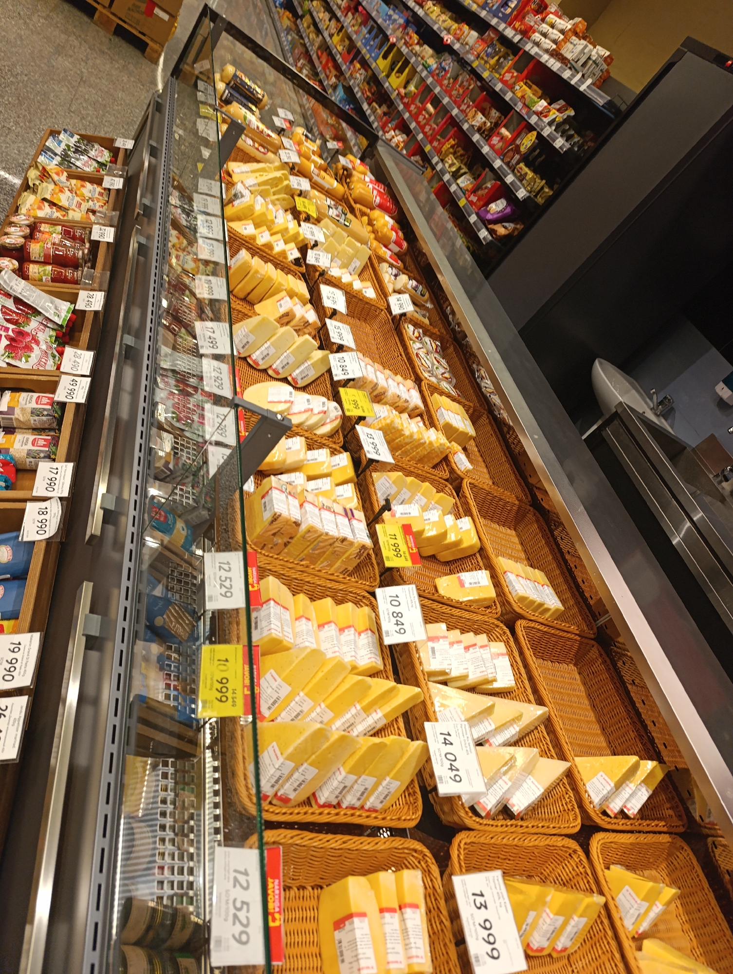 The image is a low-angle, wide shot of a refrigerated display case in a supermarket in Jizzakh, Uzbekistan. The display case is filled with various types of cheese, organized in wicker baskets and on shelves. Some of the cheese is packaged in plastic wrap, while other pieces are displayed as whole wedges or slices. Price tags are visible on many of the cheese products, with prices listed in Uzbek currency. The background shows rows of other grocery items on shelves, also in a supermarket setting. The overall lighting is bright, typical of a retail environment. There are no people visible in the image.