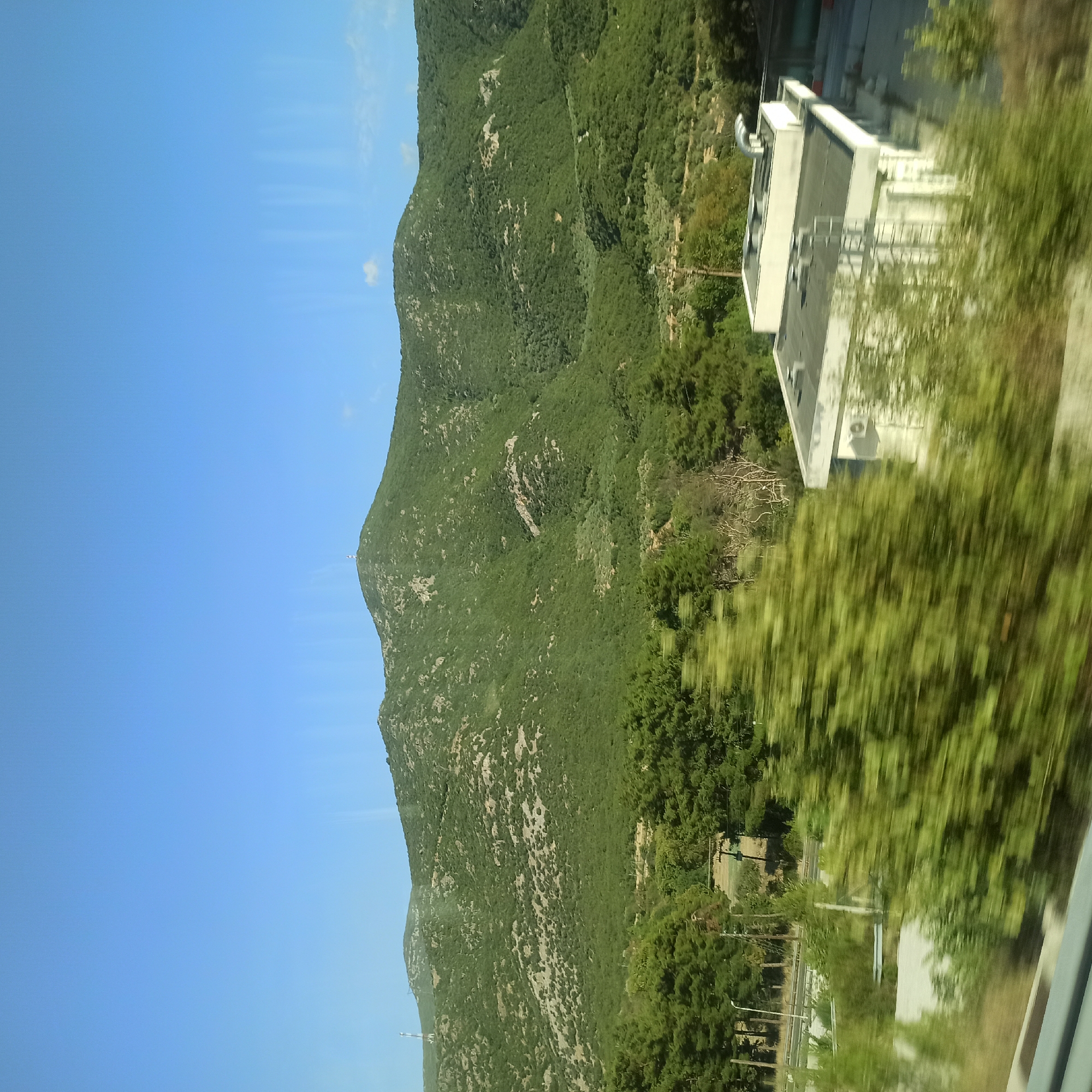 The image depicts a natural landscape with a mountainous backdrop and a clear blue sky. In the foreground, a road is visible, suggesting a view from a moving vehicle. To the right, a modern building with a flat roof and white walls is partially obscured by lush green foliage. The building appears to be a commercial or industrial structure, featuring vents and possibly an air conditioning unit. The mountains are covered in dense green vegetation, with some rocky outcrops visible. A telecommunications tower can be seen on the summit of one of the peaks. The overall scene suggests a location in a rural or semi-rural area, possibly on the outskirts of a town or city. The presence of the building and the road indicates human activity and infrastructure within this natural setting. Given the context provided, this scene is likely situated in the Municipal Unit of Ampelakia, Greece, a region known for its mountainous terrain and scenic beauty.