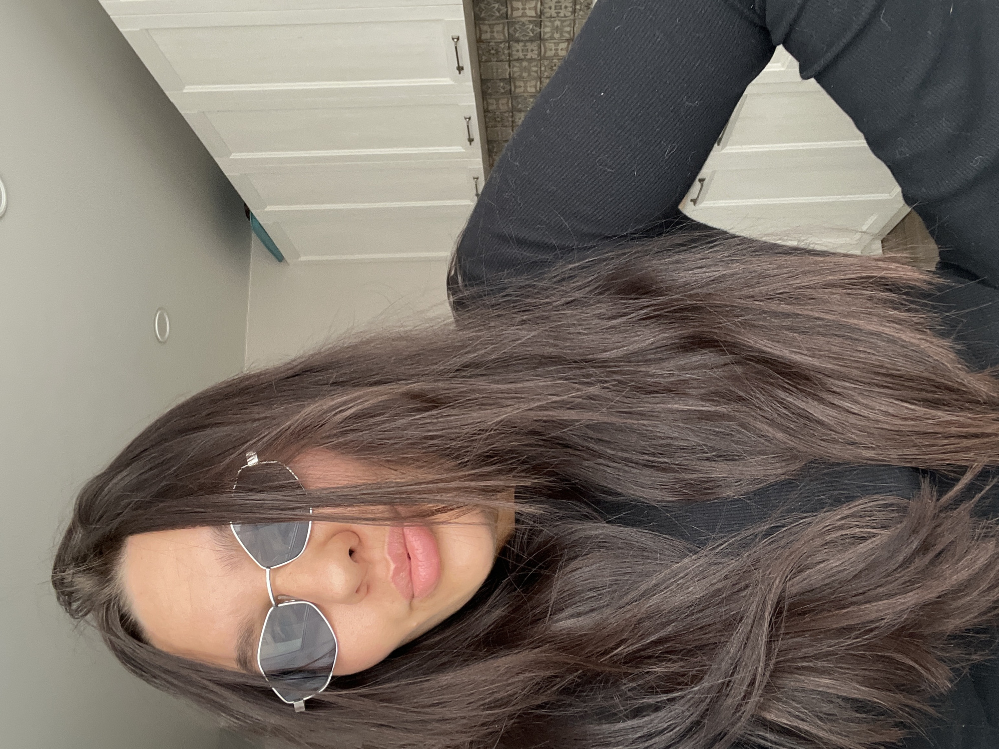 A woman with long brown hair and sunglasses is pictured indoors. She is wearing a black long-sleeved shirt. Behind her are white cabinets with silver handles and a tiled wall. The location is not discernible from the given information.