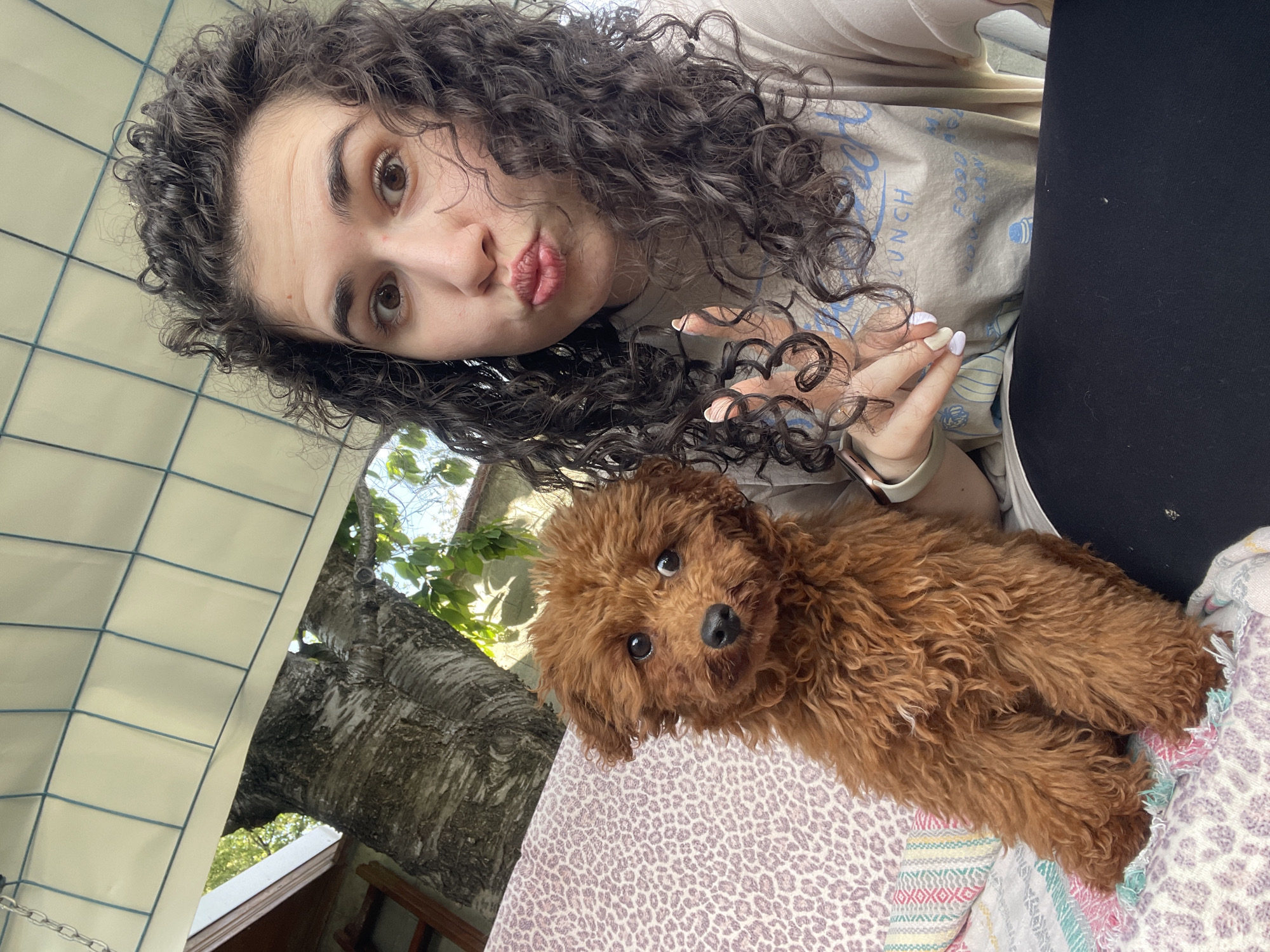 A woman with curly brown hair is taking a selfie with a brown poodle. The woman has her lips pursed and is holding up her hand. She is wearing a light-colored t-shirt. The dog is lying on a bed covered with a patterned blanket. The background shows a structure with tiled walls and a tree. The location appears to be outdoors. There is no discernible evidence to connect the picture with Sofia, Bulgaria.
