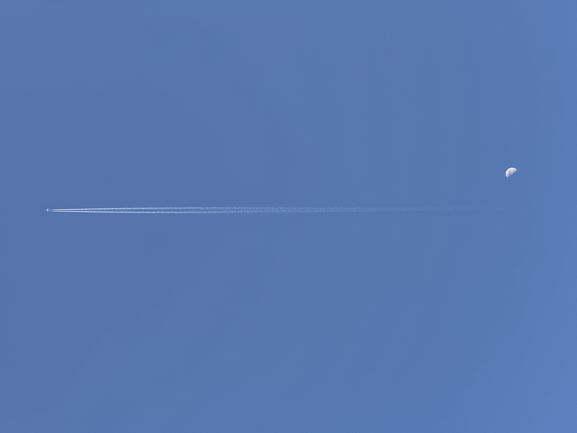 The image captures a clear, vibrant blue sky over Budva, Montenegro, during daylight hours. A white airplane is visible on the left side of the frame, flying horizontally across the sky. It leaves a long, distinct white contrail (vapor trail) that extends from the aircraft towards the right, stretching across the middle of the photograph. To the far right, a bright crescent moon is clearly visible against the blue backdrop, illuminated by the sun. The sky is entirely free of clouds, indicating calm and clear weather conditions. The scene presents a juxtaposition of an active airplane, its transient vapor trail, and the serene, distant moon, all set within the vast expanse of the daytime sky. No people are visible, and there is no discernible text within the image.