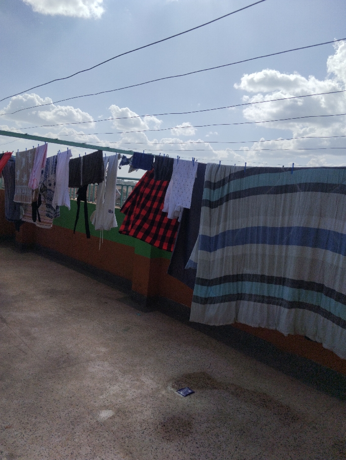 The image depicts an outdoor scene, likely a rooftop, in Witeithie ward, Kenya, on a bright day under a partly cloudy sky. Numerous garments are suspended on multiple clotheslines strung across a green metal frame. The drying laundry includes a red and black plaid shirt, various solid and patterned shirts, dark trousers, and a large striped blanket or sheet featuring blue, white, and grey/tan bands. These items are secured to the lines with blue clothespins. Below the clotheslines, a low wall or barrier is visible, painted with horizontal sections of brown and green. The ground surface is a mottled grey concrete. In the lower right foreground, a small, dark blue or purple rectangular object, possibly a wrapper or packet, rests on the ground. Overhead, several thin, dark wires traverse the blue sky, which contains scattered white cumulus clouds. No individuals are present in the scene.