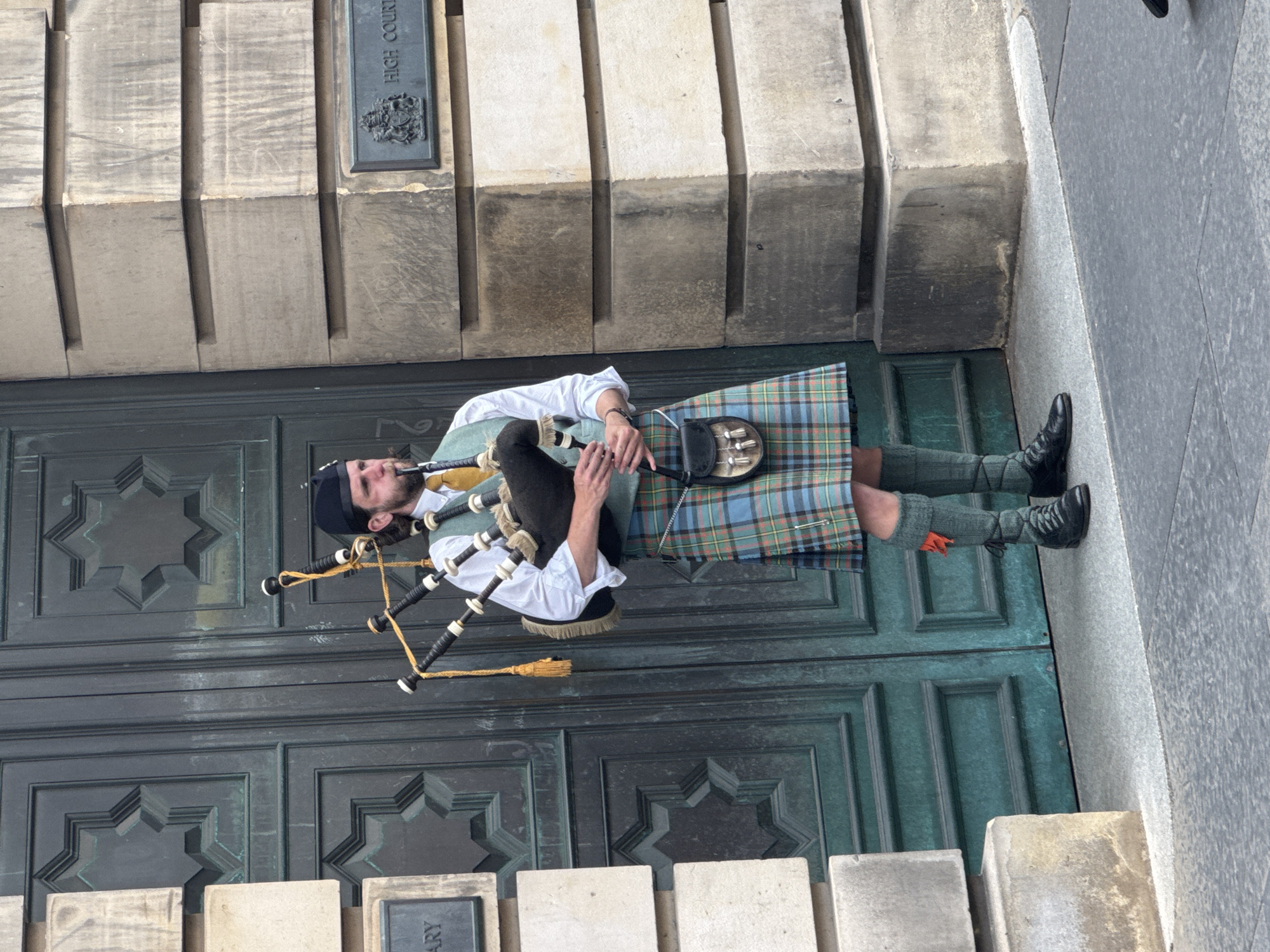 A male bagpiper stands in front of large, dark green double doors with geometric carvings. He wears a black Glengarry hat, a white shirt, a teal vest, and a kilt in a tartan pattern of green, blue, and red. He also wears green knee-high socks with orange flashes peeking out, and black shoes. He is playing the bagpipes, holding them with his right arm and blowing into the chanter with his mouth. A sporran hangs from his waist. To his right, on the stone building, is a plaque that reads "HIGH COURT". The building is constructed of large, grey stone blocks. The scene is outdoors, with a grey pavement in the foreground. This is the High Court of Justiciary on Parliament Square, City of Edinburgh, United Kingdom.