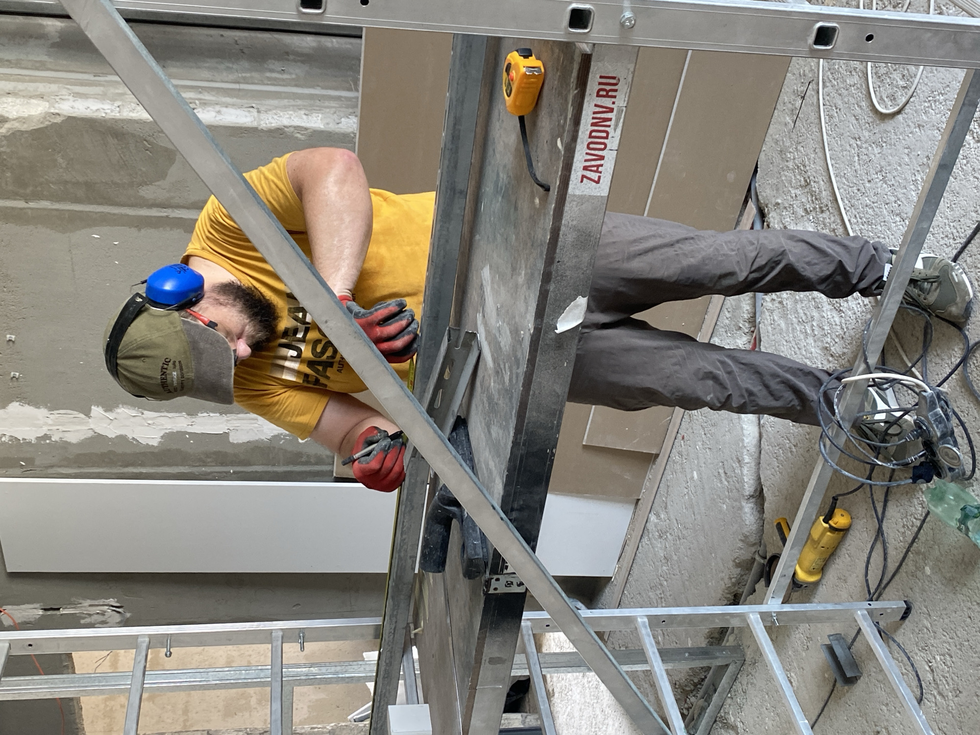 The image depicts a construction site, possibly in Batumi, Georgia, where a person is working. The individual, wearing a yellow t-shirt, brown pants, a green cap, and blue ear protection, is standing at a workbench, holding a marker and a measuring tape. They are in the process of marking a metal bar. Several tools and materials are visible, including a yellow power tool on the floor, a yellow measuring tape on the workbench, and various metal components. The environment appears to be an indoor construction area with unfinished walls and debris on the floor. A ladder is also present in the scene.