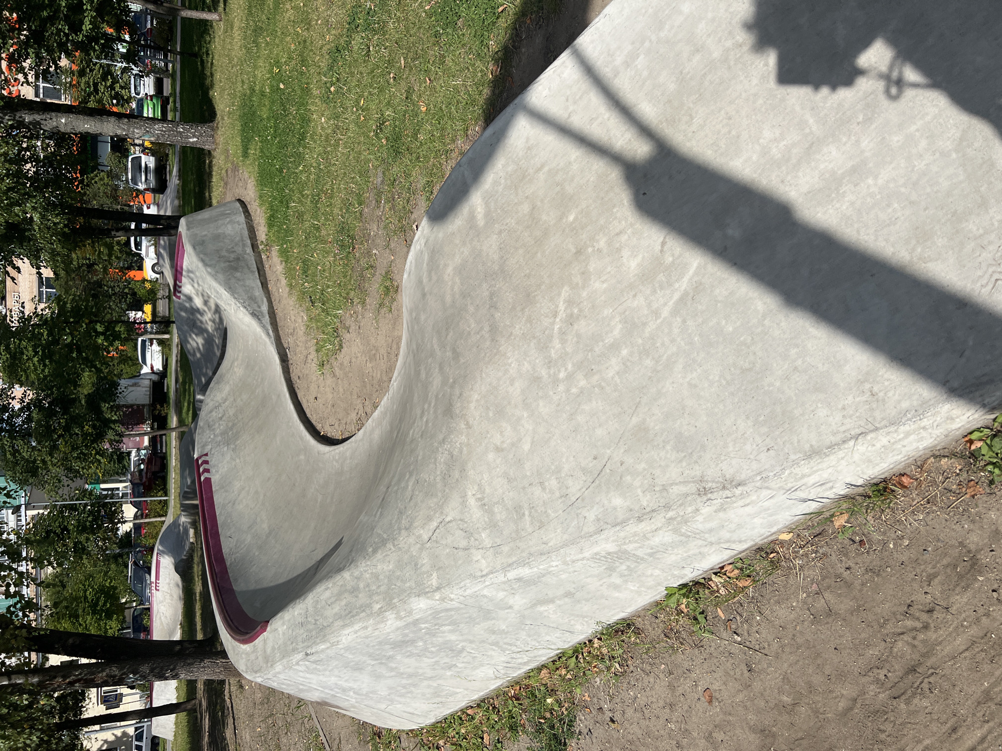 A low-angle, wide shot captures a concrete pump track in a park-like urban setting. The track is a series of smooth, undulating curves made of light gray concrete, with a darker, reddish-purple stripe along the top edges of some sections. The track is positioned in the foreground and midground, curving from left to right across the frame. Behind the track, in the background, are trees with green foliage, parked cars, and buildings. A shadow of a person holding a phone, presumably taking the photograph, is cast across the concrete track in the lower right portion of the image. The ground surrounding the track is a mixture of dirt and sparse grass. The scene appears to be set in a city park or recreational area within Moscow, Russia, evidenced by the architecture and the presence of a common urban park design.