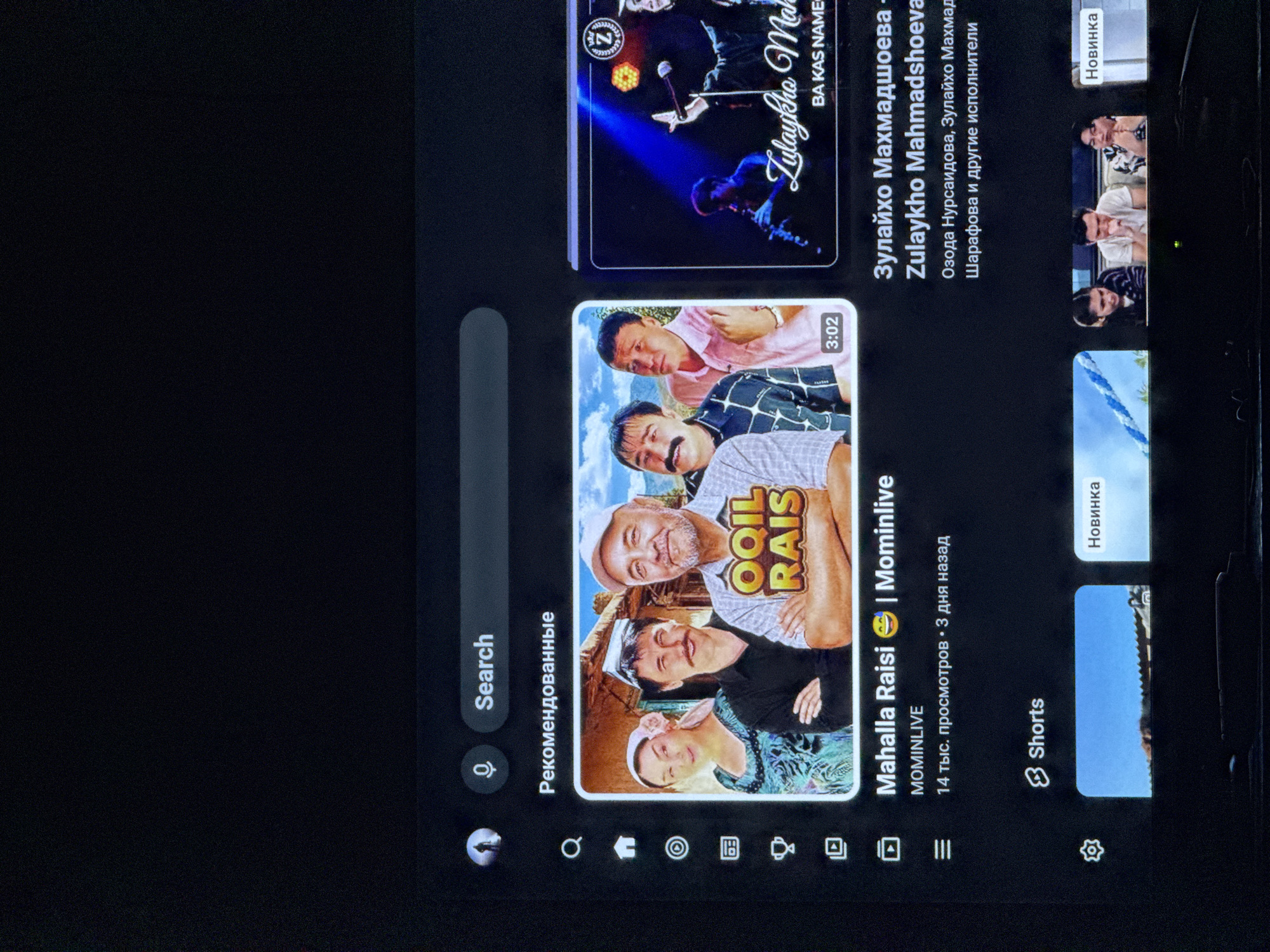 This is a media file that appears to be a screenshot of a YouTube-like interface, likely on a mobile device or tablet, given the aspect ratio and interface elements. The content displayed is a mix of video thumbnails and interface options.
The main subject is a collection of video thumbnails featuring people, predominantly of Central Asian descent, dressed in traditional attire. One prominent thumbnail shows three individuals, two men and a woman, with the text "OOIL RAIS" overlaid. Another thumbnail features a performer on a stage with vibrant lighting, accompanied by Cyrillic text that translates to "Zulaykha Mahmadshoeva". There are also thumbnails labeled "Shorts" and "Новинка" (New).
The interface elements include a search bar with a microphone icon, a "Search" label, and "Рекомендованные" (Recommended). At the bottom, there's a navigation bar with icons suggesting options like home, search, subscriptions, library, and settings.
The overall scene suggests a user browsing or interacting with video content, possibly entertainment or music. The presence of Cyrillic text and the appearance of the individuals strongly suggest a context related to Uzbekistan, and the prompt specifies Bukhara, Uzbekistan, as the location. The images evoke a sense of cultural performance and everyday life. There are no explicit indicators of time of day or weather. The emotions conveyed by the individuals in the thumbnails range from smiling to more neutral expressions.