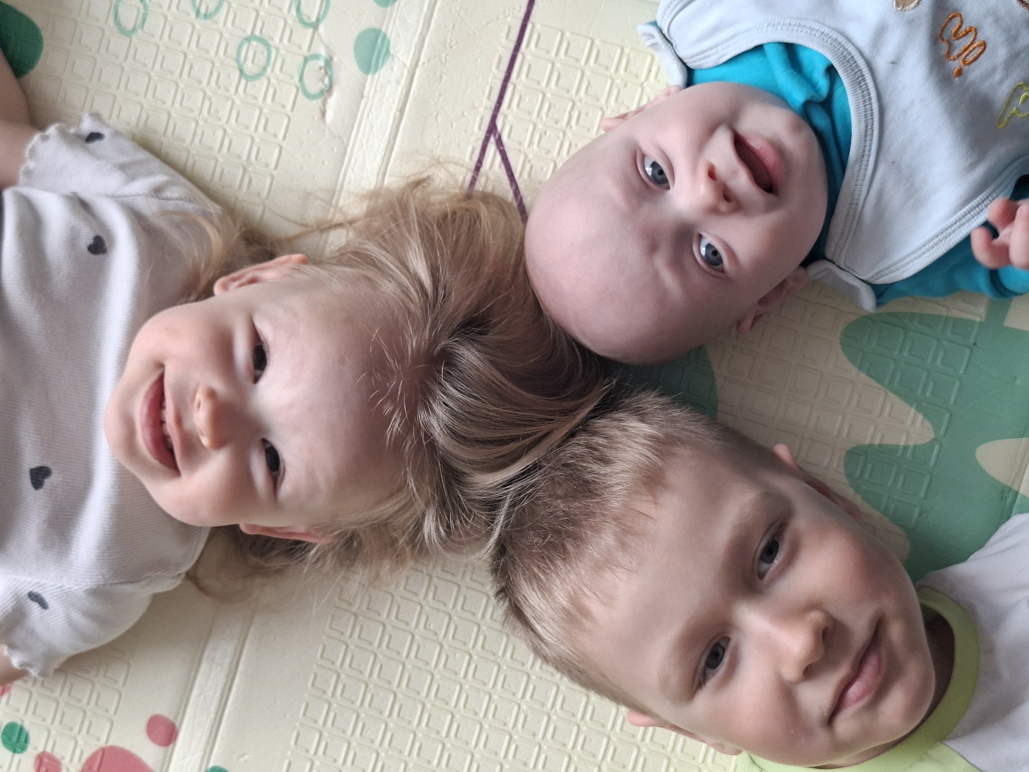 Here is a forensic description of the image.
Three children lie on a colorful, cushioned mat. A toddler in a white shirt with small black hearts smiles towards the viewer, her blonde hair spread out on the mat. Above her, a baby wearing a light blue onesie with a blue undershirt also looks at the viewer, smiling with an open mouth. To the right and below the toddler, a young boy in a white shirt with a green collar looks slightly off to the side. The mat they are lying on has a grid pattern with colorful circles and shapes.