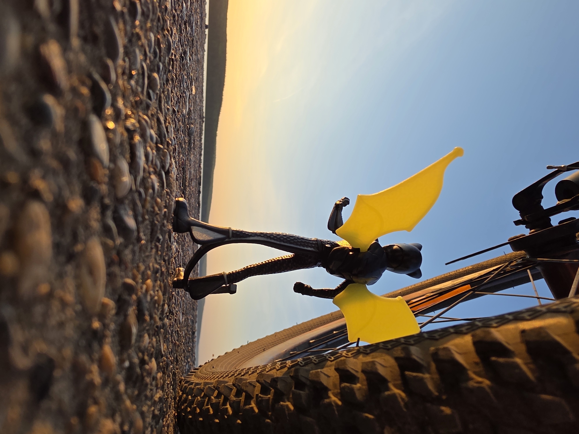 A low-angle, slightly tilted shot depicts a scene from the perspective of someone on a bicycle at what appears to be the Kama Reservoir in Chaikovsky, Russia.
Foreground: A bicycle tire with a knobby tread is prominent. Perched on the handlebars is a black, skeletal figure with yellow wings attached.
Midground: A pebbled shoreline stretches along the left side of the frame.
Background: A sliver of land is visible on the horizon, illuminated by the setting sun. The sky transitions from a golden hue near the horizon to a light blue overhead.